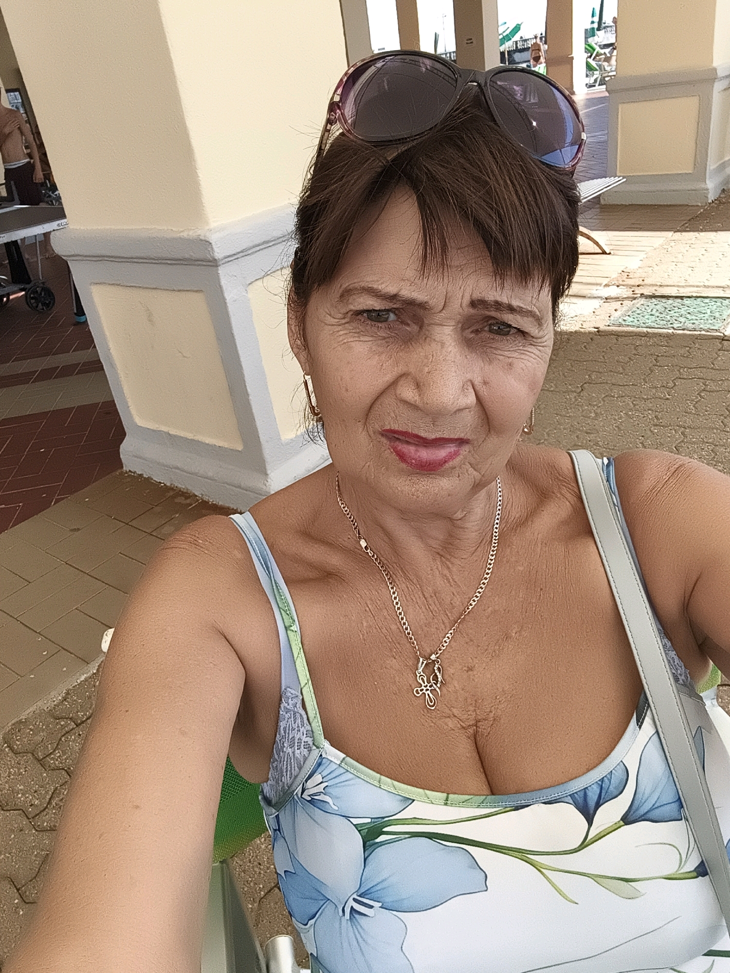 A woman is taking a selfie. She is wearing a floral print top, a gold necklace, and large sunglasses on her head. She has brown hair and appears to be outdoors, possibly in a public area with light-colored pillars and patterned flooring. The background suggests a sunny day, with some people and beach chairs visible in the distance. There are no specific elements in the image that would indicate a location in Livorno, Italy.