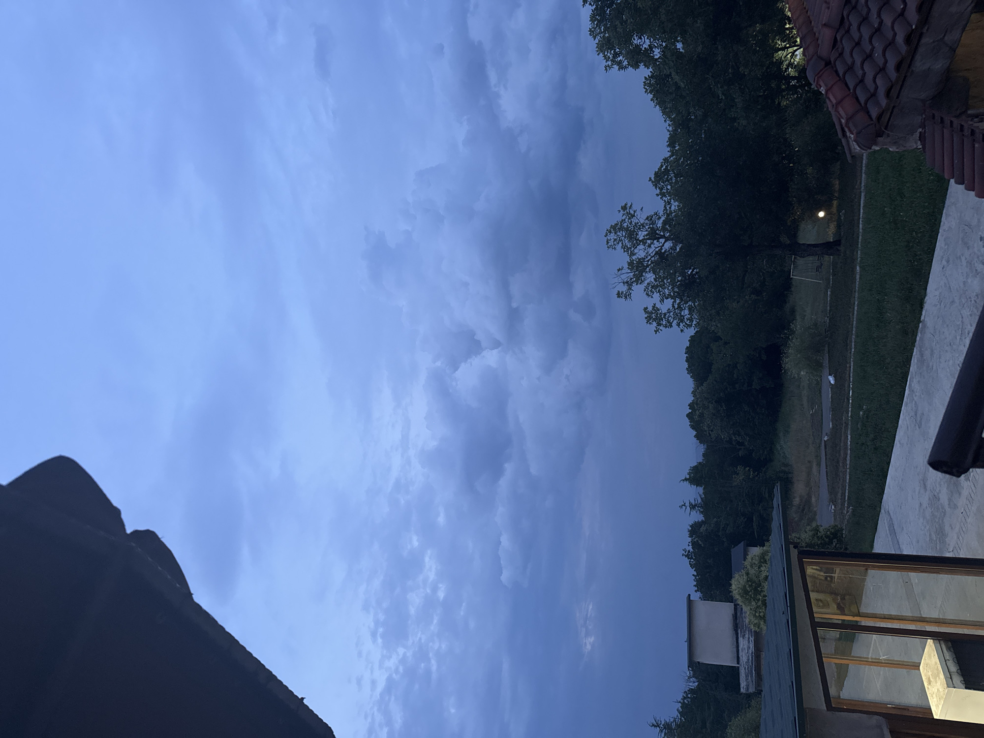 The image depicts a cloudy sky over a semi-rural or suburban landscape, likely in or around Sredets, Bulgaria, given the provided context. In the foreground, the dark eaves of a building are visible, casting a shadow. To the left, a portion of a modern building with large glass windows and a green roof is present. Beyond this structure, the landscape transitions to a grassy area with some scattered trees and a small building with a light-colored rectangular structure on its roof. Further in the background, a dense line of dark green trees forms a natural boundary. In the middle ground, a solitary lamp post with a lit bulb illuminates a patch of grass beneath a large, dark-leaved tree. There is also a swing set visible near the tree. The sky is filled with dramatic, dark clouds, suggesting either dawn, dusk, or an approaching storm. No people are visible in the frame.