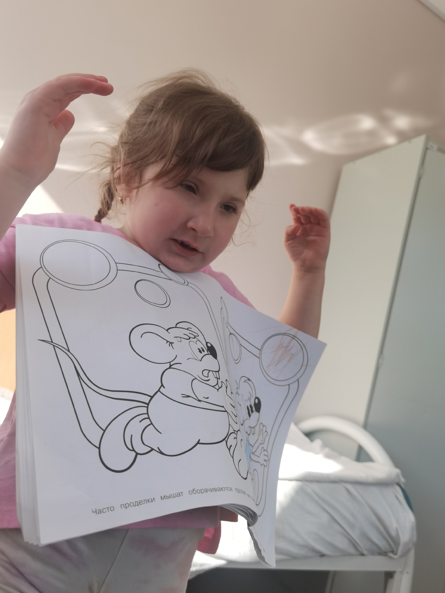 A young girl with light brown hair and a pink shirt is holding a coloring book in front of her chest. Her hands are raised in the air, palms facing inward. The coloring book features a cartoon image of two mice and some Russian text. The text translates to "Often the antics of the mice turn against them." The scene appears to be indoors, possibly in a room in Perm, Russia, based on the Russian text in the coloring book. The background shows a white cabinet, a hospital bed covered with white linens, and a white wall.