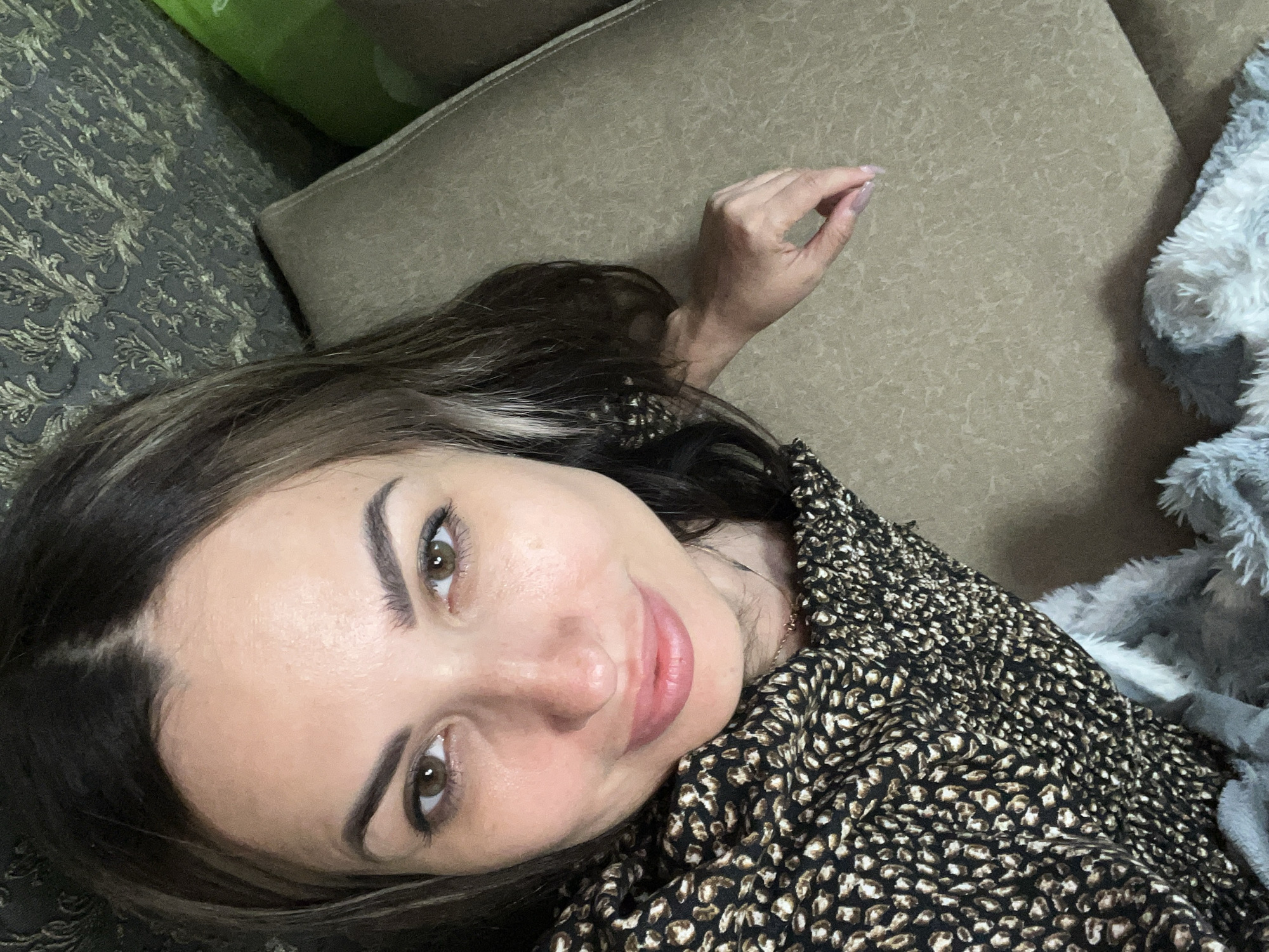 A woman with long, dark hair with blonde highlights is lying on a couch. She is looking directly at the camera and smiling. She is wearing a black and brown leopard print top. Her left arm is resting on the couch, and her hand is visible. There are pillows and a blanket on the couch with her. The background is a patterned wall and a green pillow. The image was taken indoors.