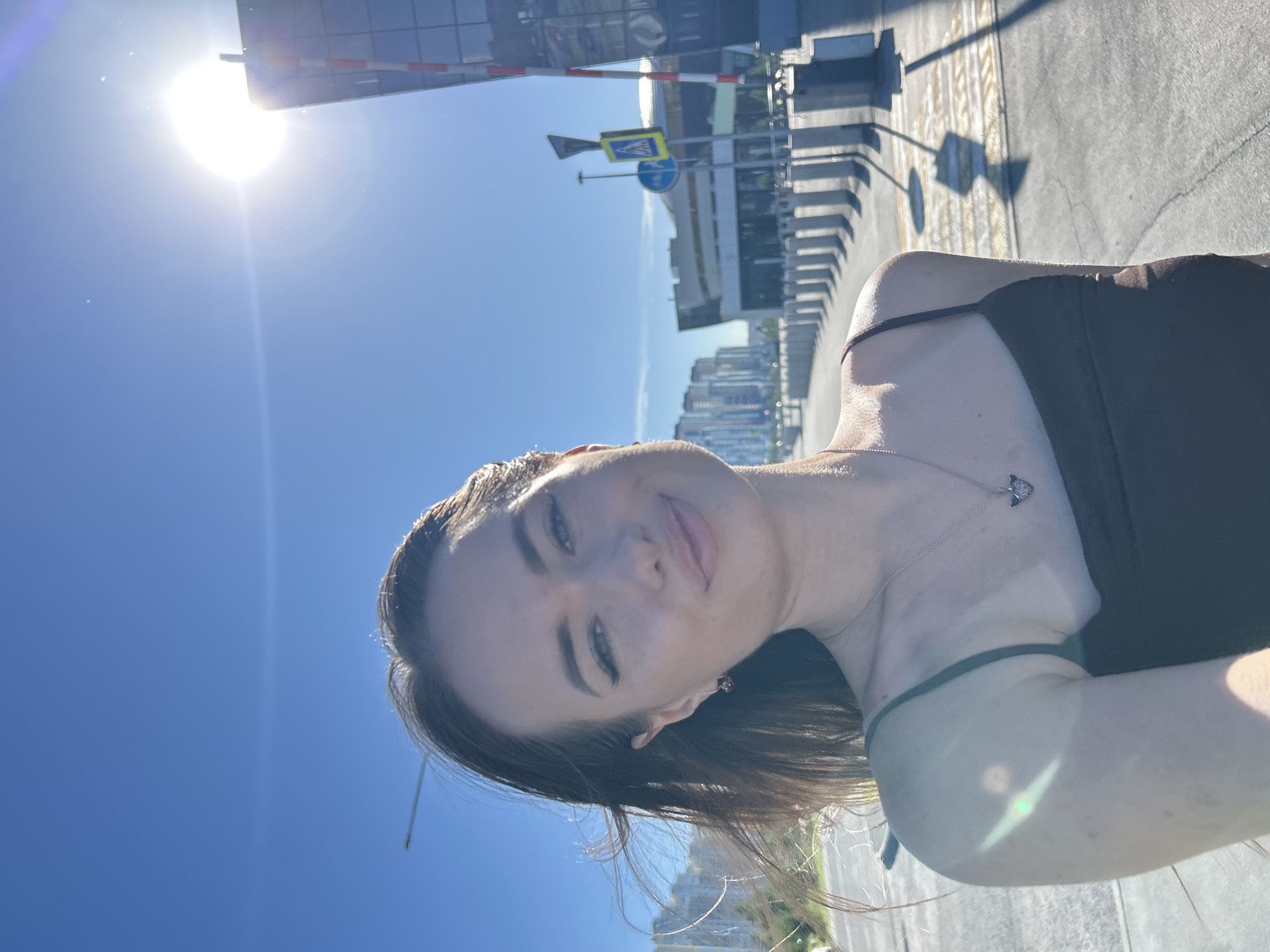 The media file shows a young woman taking a selfie outdoors on a bright, sunny day. The sun is high in the sky, casting strong shadows. She is wearing a dark tank top and a delicate necklace. Her hair is dark and appears to be blowing slightly in the breeze. The background shows a modern urban environment with several tall buildings and a road with painted lines. There are also traffic signs visible, including a pedestrian crossing sign. The overall mood seems casual and relaxed. It is daytime, likely late morning or early afternoon given the strong sunlight. The woman's expression is a slight smile, suggesting she is enjoying the day.