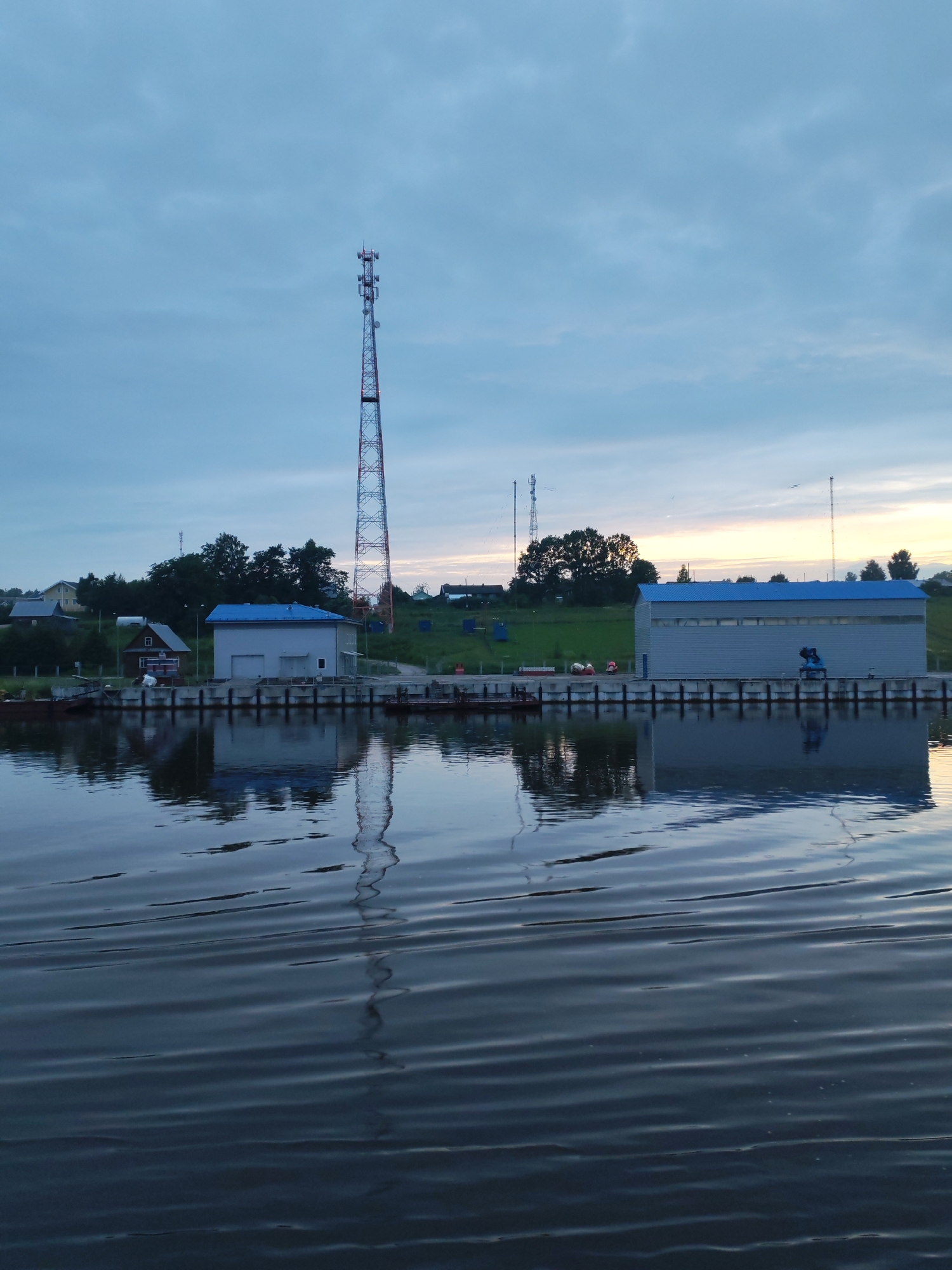 The image depicts a tranquil scene at the edge of a body of water, likely a river or canal, in Ivanov Bor, Russia. The time appears to be dusk, as indicated by the soft, diffused light and the muted colors of the sky. In the foreground, the calm water reflects the structures on the shore and the sky above, creating a mirror-like effect. The water is rippled, suggesting a gentle breeze or the wake of a passing boat.
Along the shoreline, a concrete quay or embankment is visible, lined with what appear to be pilings or mooring posts. Several buildings are situated along this embankment. The most prominent is a long, modern-looking industrial or warehouse-style building with a blue, corrugated metal roof and grey, ribbed metal walls. To the left of this building is a smaller, older structure with a white facade and a dark-colored roof, possibly a house or an office. Further to the left, partially obscured by trees and a gentle slope of green grass, are several other residential-style houses with pitched roofs, suggesting a small settlement or village.
Dominating the skyline and serving as a focal point is a tall, lattice-style transmission tower, likely for telecommunications or broadcasting. Its red and white sections are clearly visible against the pale blue and grey sky. Two other, shorter towers, also appearing to be transmission masts, are visible further in the background. The sky itself is a blend of soft blues and greys, with hints of warmer tones near the horizon, suggesting the last vestiges of sunset. The overall impression is one of a quiet, functional industrial area situated within a natural, possibly rural, setting. There are no people or animals visible in the frame.