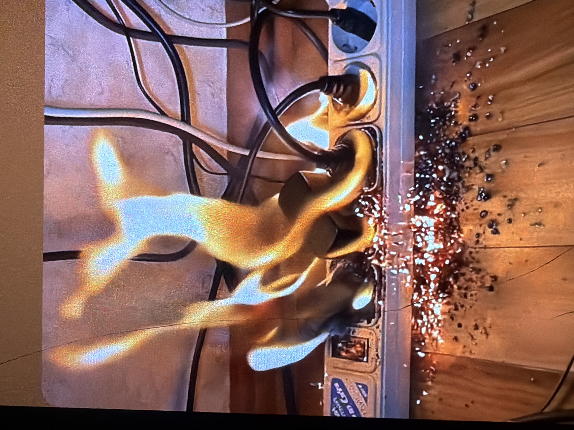 The image shows a close-up of a power strip that is on fire. Flames are erupting from multiple outlets, with electrical cords and plugs visible. Sparks and burning debris are scattered on the wooden surface below the power strip. The background consists of a textured wall and more electrical cords. The scene depicts a hazardous electrical fire. No specific location in Bukuru, Nigeria can be determined from the image.