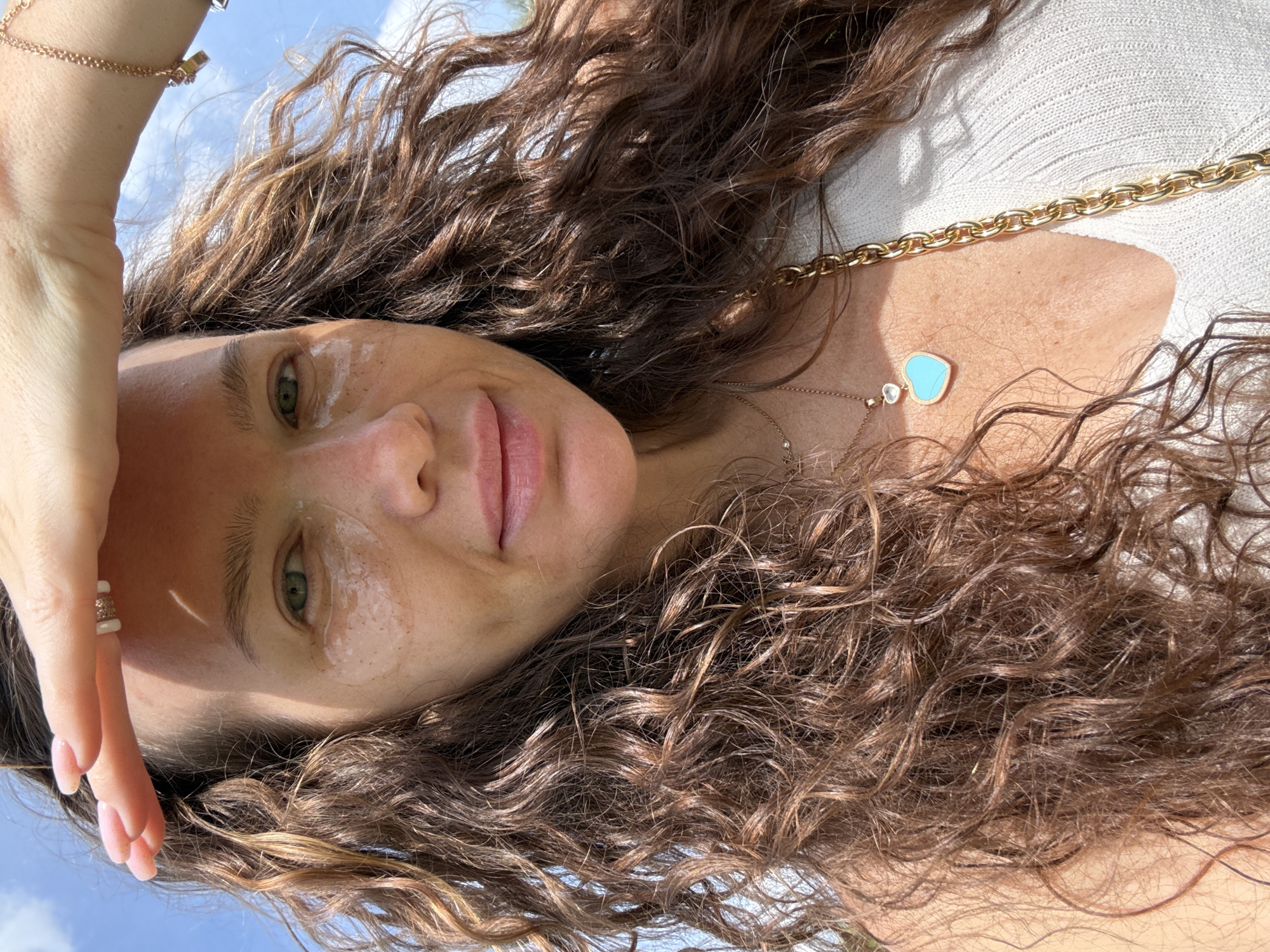 A woman is looking up at the sky, shielding her eyes with her hand. She has long, curly brown hair and is wearing a white top and gold jewelry, including a necklace with a blue heart-shaped pendant. She has under-eye patches on her face. The background is a bright blue sky with some clouds. The scene appears to be outdoors on a sunny day. No specific location in Moscow, Russia can be determined from the image.