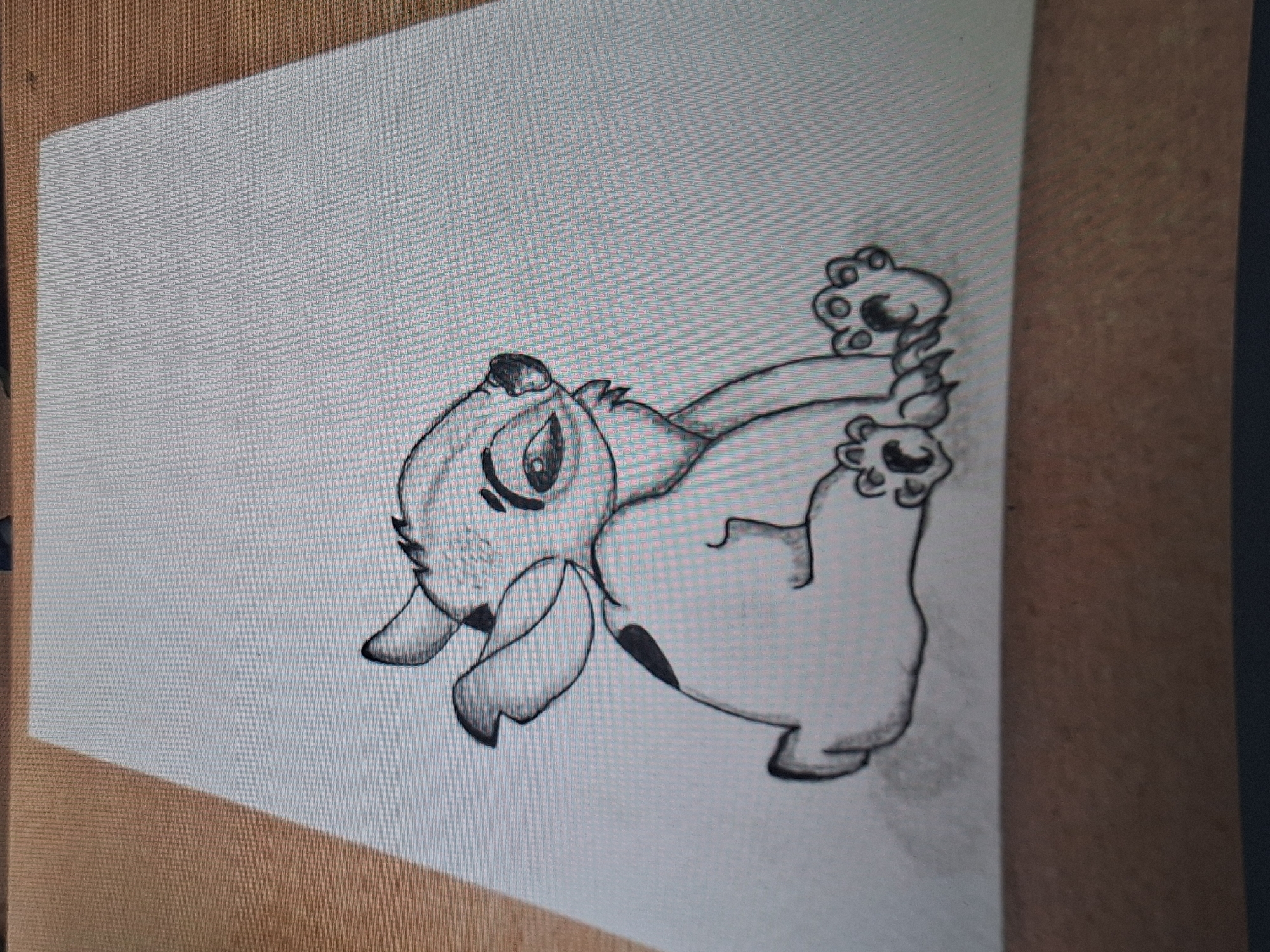 The image displays a hand-drawn sketch of a character resembling Stitch from Disney's Lilo & Stitch, rendered in black pencil or charcoal on a light-colored, rectangular sheet of paper. The character is depicted in a seated posture, turned slightly to its right, with its head tilted and gaze directed towards the right side of the frame. Key features include large, drooping ears, prominent oval eyes with dark pupils, a small nose, and a subtle, somewhat pensive or melancholic mouth line. Two distinct, dark, irregular spots are visible on its back or upper left arm. Its front and hind paws are detailed with visible pads and claws. The drawing incorporates shading to add depth and texture around the face, ears, and limbs, and a light shadow is cast beneath the character on the paper surface. The paper itself exhibits a fine, grid-like texture, likely originating from a digital display or the paper's weave. The drawing rests on a flat, light-brown wooden surface, which displays a visible wood grain pattern, consistent with a table or desk. The lighting suggests an ambient source from the upper right, creating a faint shadow along the top-left edge of the paper. No visual information within the image identifies a specific geographic location, including the city of Varna, Bulgaria.