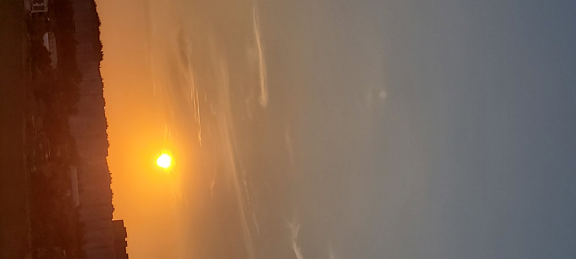 A vertical, low-angle shot captures a sunset over a city skyline. The sky transitions from a light blue at the top to a warm orange near the horizon. Wispy clouds streak across the sky. The sun, a bright white-yellow disc, is positioned in the upper right quadrant of the image. Below the sky, a silhouetted city stretches horizontally, with rows of apartment buildings and trees forming a dense urban landscape. The bottom of the image shows a darker, indistinct foreground. The overall impression is of an urban setting bathed in the warm light of dusk.