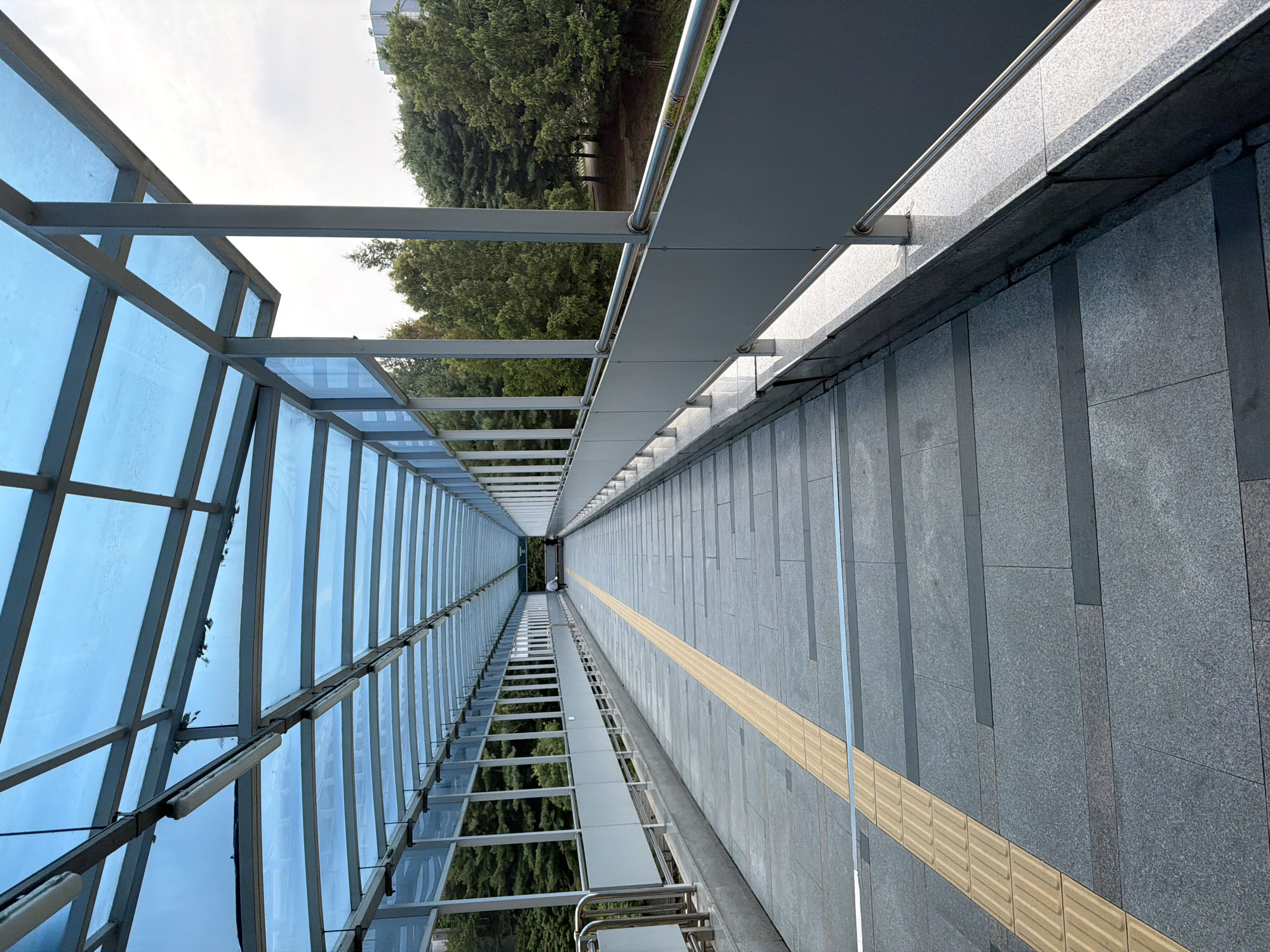 The image depicts a long, covered pedestrian walkway with a blue, translucent roof supported by a metal frame. The walkway is paved with large, grey granite tiles. A tactile paving strip, likely for the visually impaired, runs along the left side of the walkway. The walkway is bordered by a metal railing and a solid grey panel on the right, and a white railing and panel on the left. Trees and foliage are visible beyond the left railing, suggesting an outdoor, possibly park-like setting. In the distance, at the end of the walkway, a lone figure in white is walking away from the camera. Another person is partially visible further down the walkway. The walkway appears to be elevated, as indicated by the presence of supports below the left side. The overall impression is one of a functional, modern public space. This scene is likely located in Uijeongbu-si, South Korea, given the context provided.