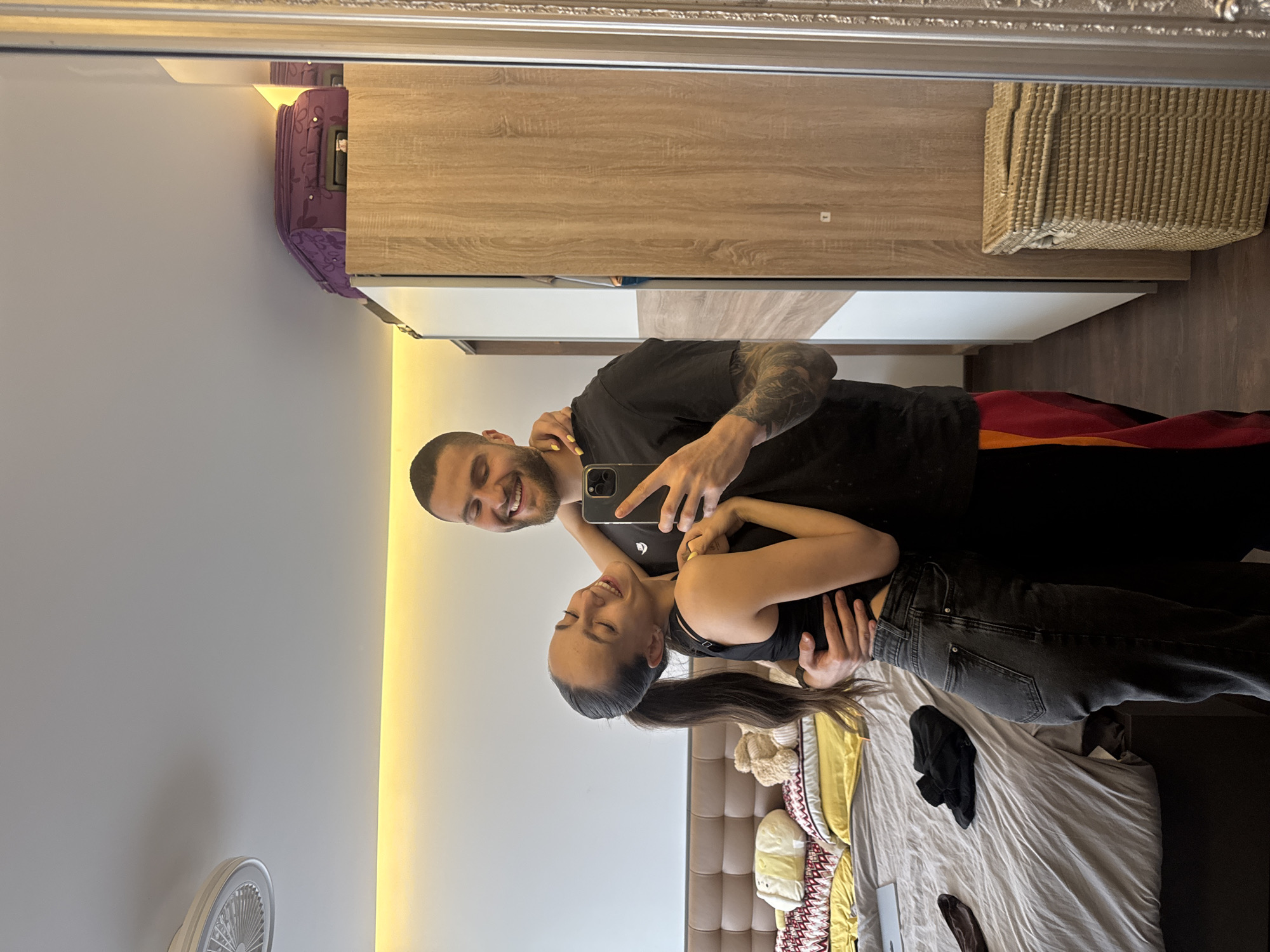 A man and woman are reflected in a mirror. The man, on the left, is taking a selfie. He has short dark hair and a beard and is wearing a black t-shirt with a tattoo on his left arm. The woman is leaning into the man and smiling. She has long brown hair pulled back in a ponytail. She is wearing a black tank top and gray jeans. They appear to be in a bedroom with a bed, pillows, and blankets visible in the reflection. A suitcase and wicker basket are also in the image. Given the style of furniture and decor, it is likely this is a modern apartment. The exact location in Sofia, Bulgaria, cannot be determined from the image.