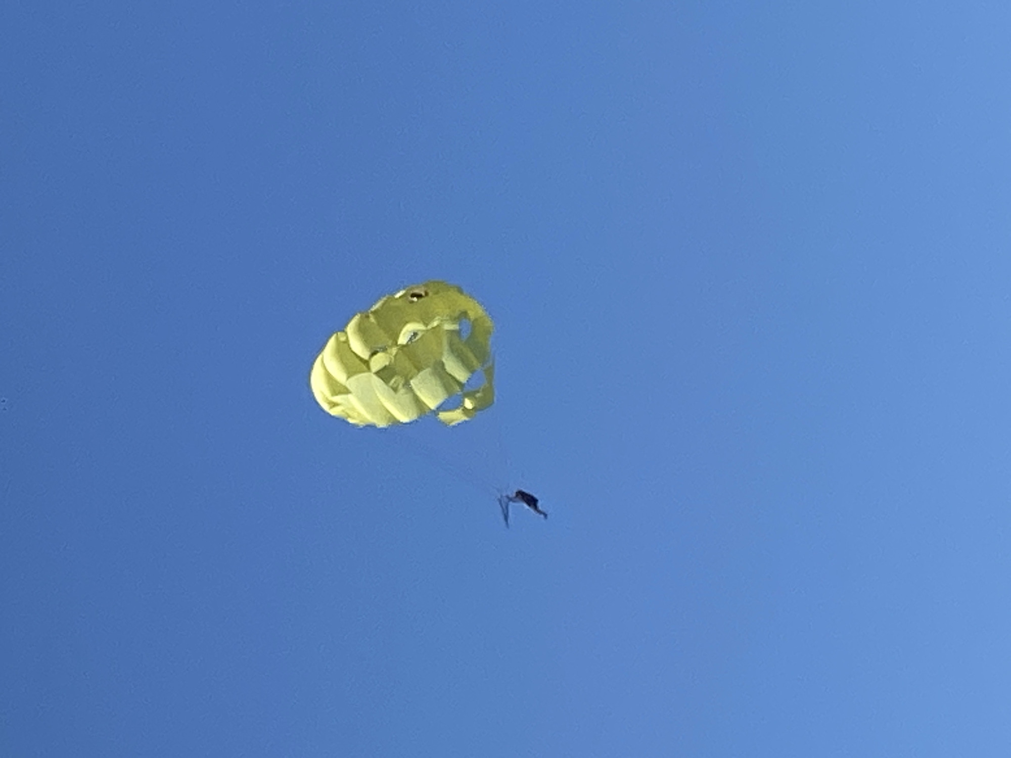A low-angle, distant shot shows a person parasailing on a partly sunny day in Alanya, Turkey. The sky is a cloudless blue. The parachute is pale yellow, and the parasailor, in a black suit, is suspended beneath it by cables.
