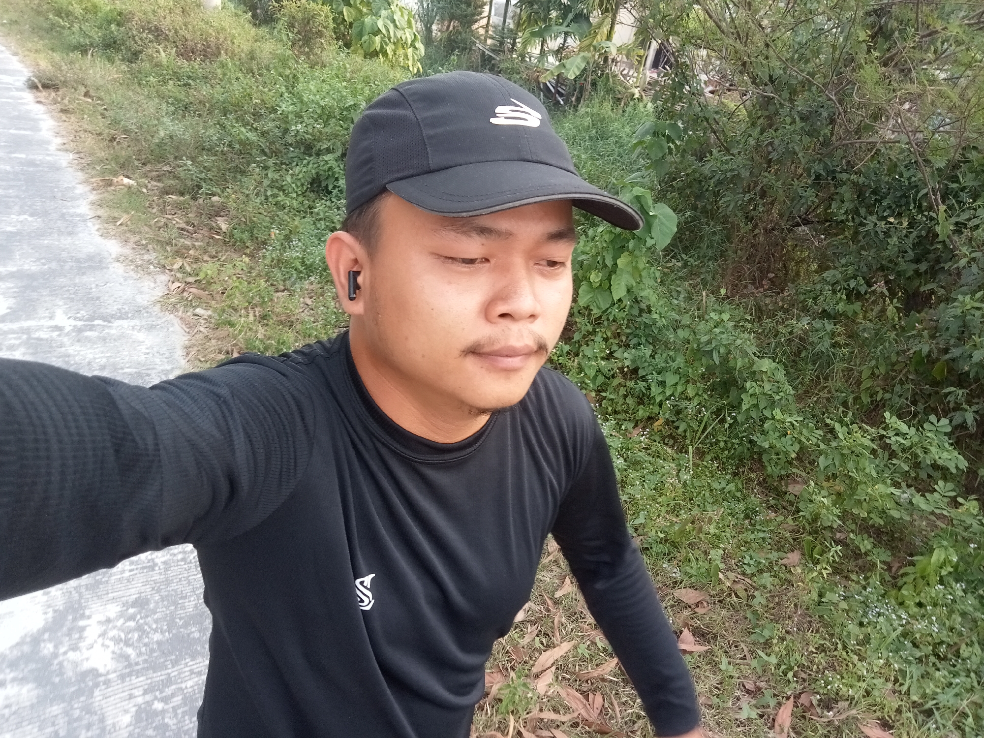 A man wearing a black baseball cap and a black long-sleeved shirt is visible in a selfie taken outdoors. He has a mustache and is wearing a black wireless earbud in his left ear. The background consists of lush green foliage and a paved path. The scene appears to be in a rural or natural setting, likely within the city of Teluk Kenidai, Indonesia. The lighting suggests it is daytime, with soft, natural light.