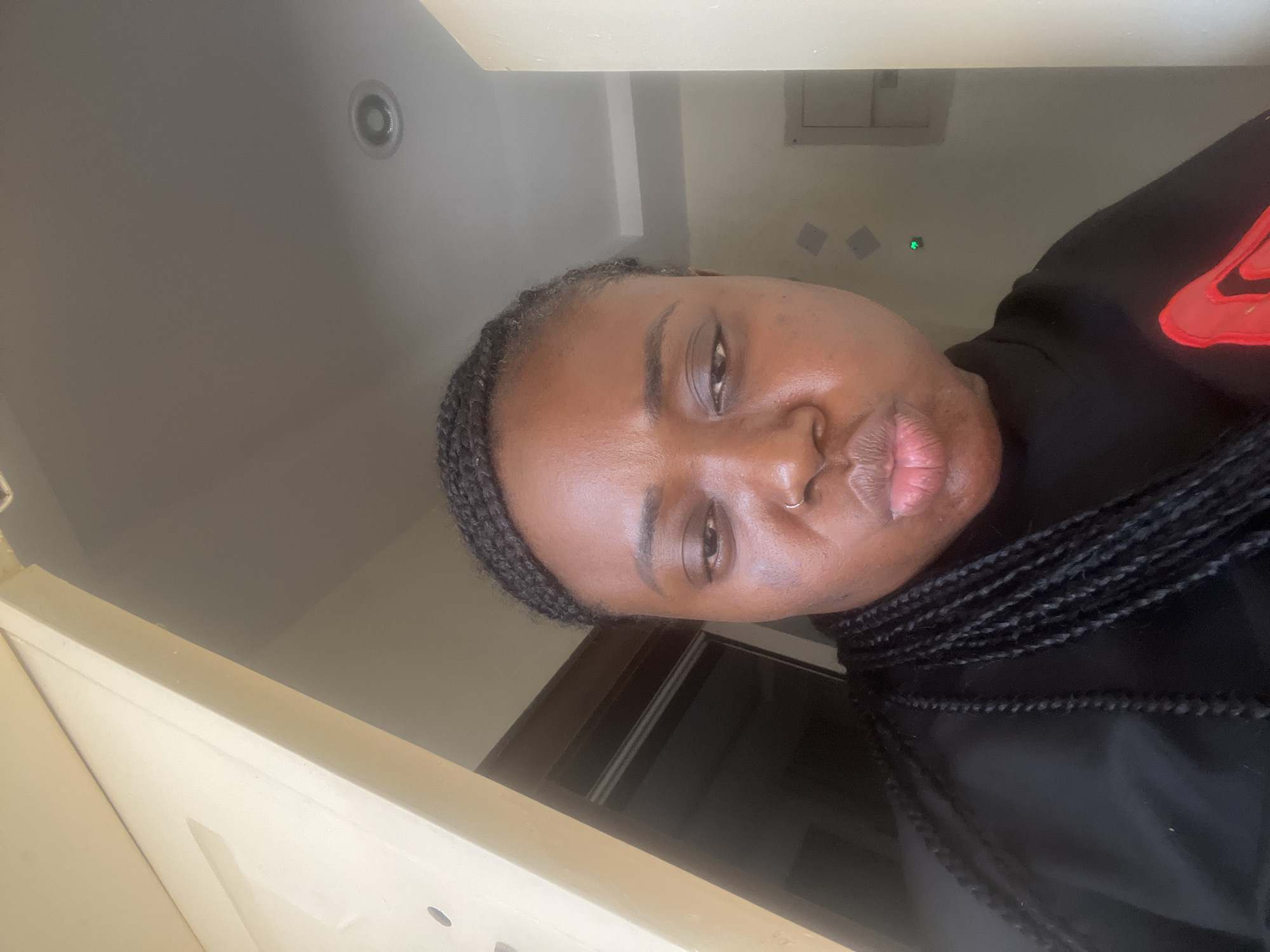 The image is a vertical, close-up selfie of a Black individual, likely female, taken indoors in Jos, Nigeria. The person is looking directly at the camera with a neutral to slightly pouting expression. They have dark skin, brown eyes, and a small silver nose ring on their left nostril. Their hair is styled in dark box braids, visible around their face and extending over their shoulders.
They are wearing a black long-sleeved top or sweatshirt, which features a prominent red logo on the right side of their chest (viewer's left), resembling the Nike swoosh.
The background consists of light-colored walls and a ceiling. A circular recessed light fixture is visible on the ceiling in the upper right. On the wall behind the person, a small, faint green light dot is present, possibly an indicator light from an electronic device. Subtle rectangular shapes can also be discerned on the wall. A light-colored doorframe or wall section is partially visible in the lower left foreground. The lighting appears to be ambient indoor light during the daytime.