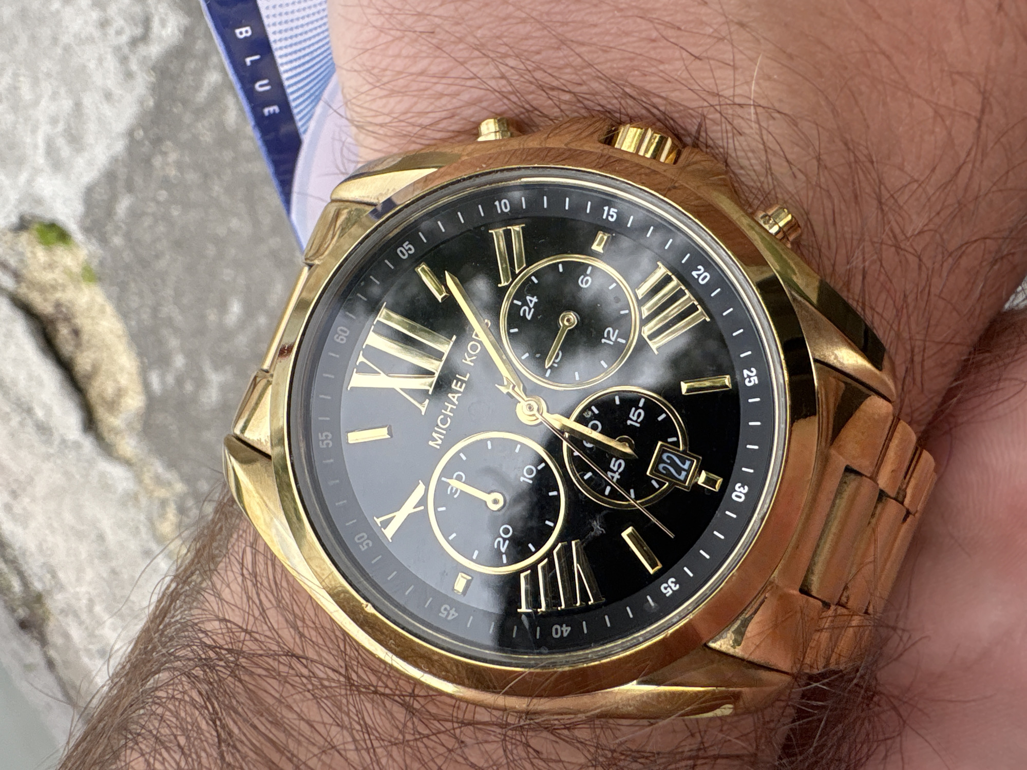 The image shows a gold Michael Kors chronograph watch on a person's wrist. The watch has a black face with gold Roman numerals and hands, and three subdials. The date window displays "22." A piece of blue fabric with the word "BLUE" printed on it is visible near the watch. The arm is quite hairy. The background appears to be an outdoor setting, possibly a sidewalk or pavement. The information available does not permit ascertaining the location of Yerevan, Armenia.