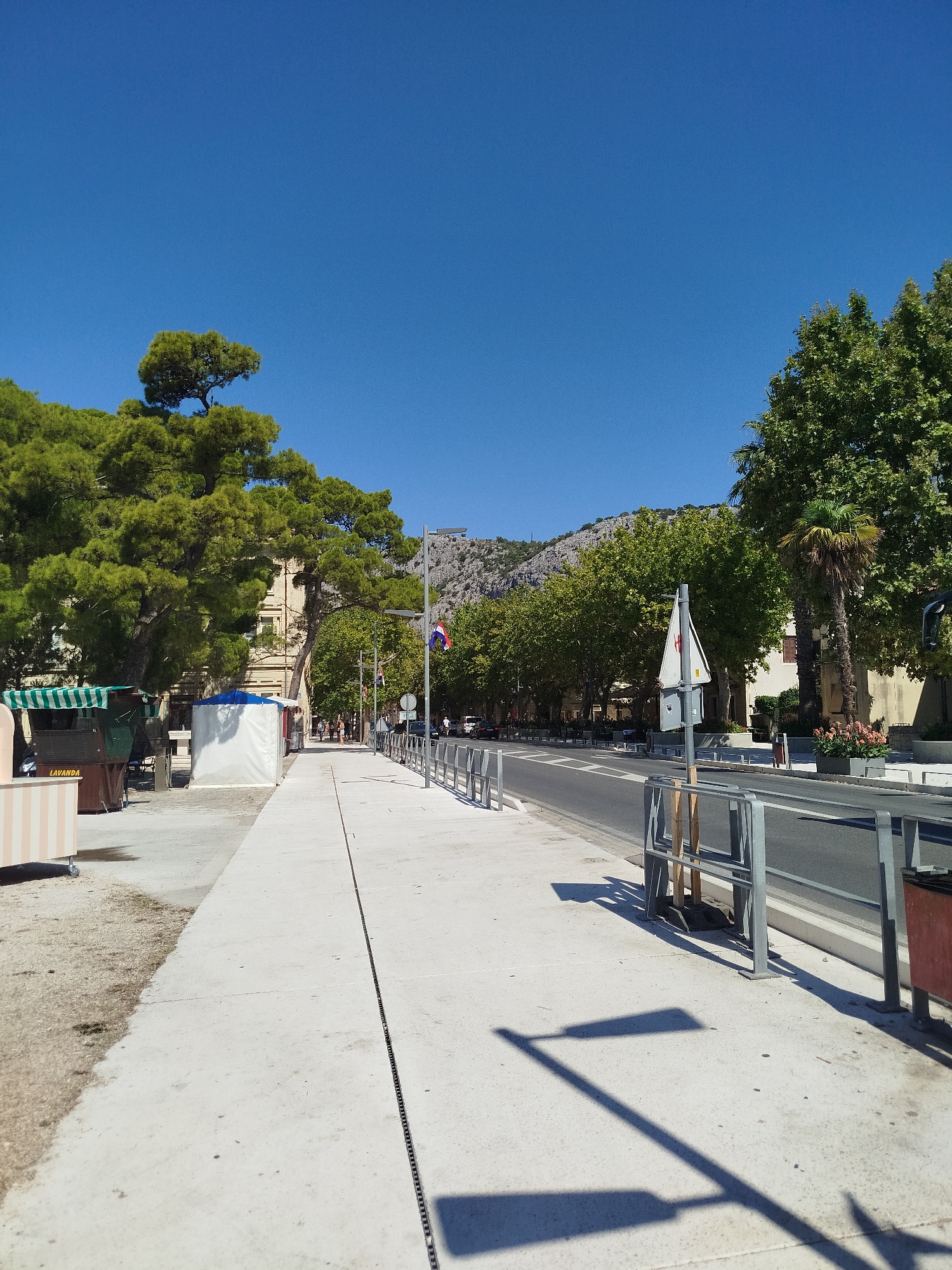 A straight, wide, paved sidewalk runs diagonally from the lower left to the upper right of the image, bordered by a metal railing on the right and a line of trees on the left. Beyond the railing is a two-lane road with parked cars and moving traffic. The sidewalk itself is clean and well-maintained, with a dark seam running down its center. To the left of the sidewalk are several market stalls with striped awnings and a large pine tree with lush green foliage. In the background, a building with light-colored walls and windows is visible, along with more trees and a mountainous landscape. The sky is a clear, bright blue, indicating a sunny day. The scene appears to be in a coastal city, possibly a promenade or a street lined with shops and cafes. The presence of market stalls and the architectural style of the building suggest a Mediterranean location. The Croatian flag is visible on a flagpole to the right of the road. The overall atmosphere is one of a pleasant, sunny day in a vibrant urban environment.