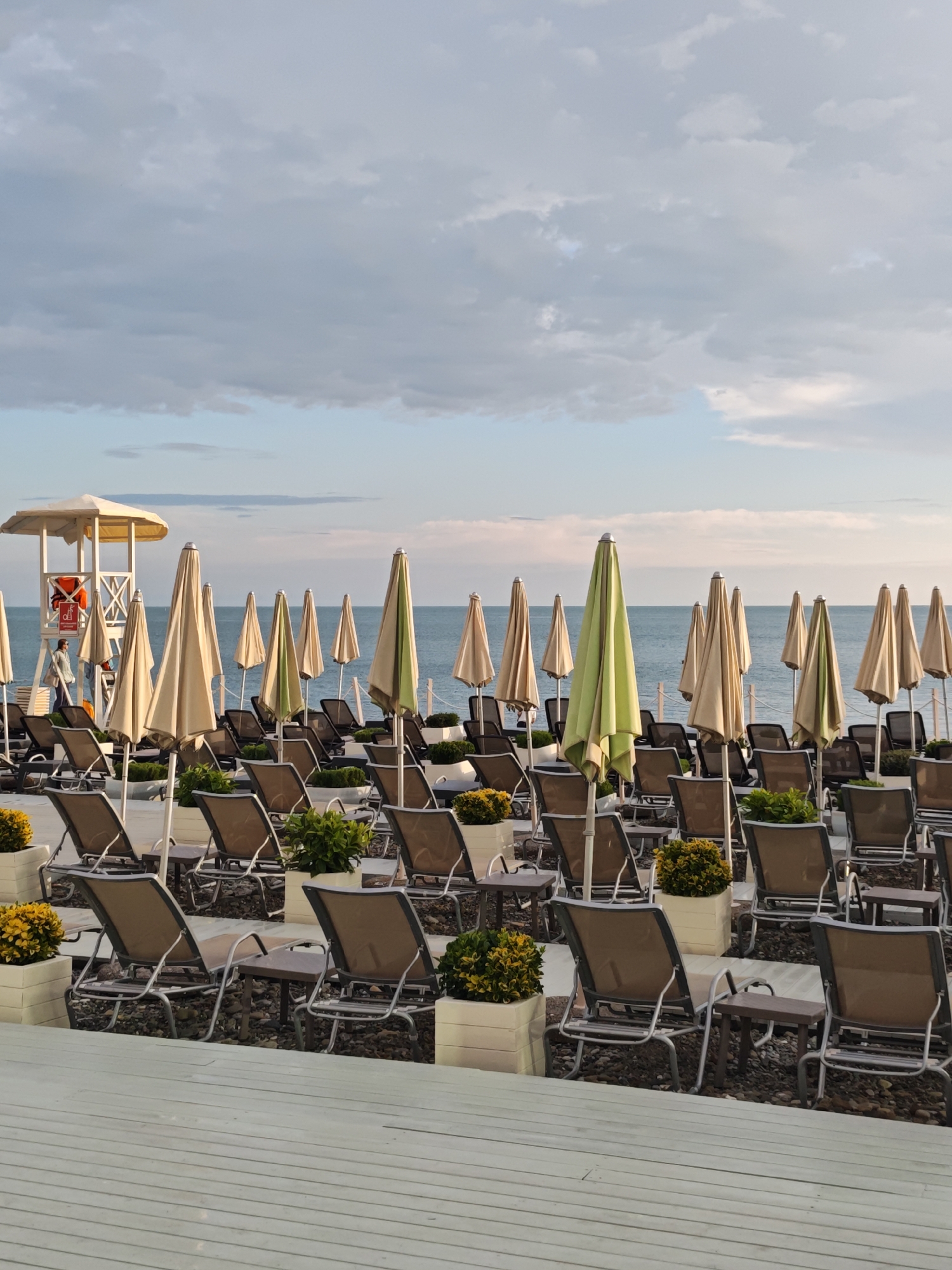 The image shows an outdoor beach setting with rows of empty lounge chairs and closed umbrellas in the foreground. Each pair of chairs has a small table between them, and planters with greenery decorate the rows. In the background is a lifeguard station, with a person walking near it. Beyond the chairs, the sea is visible, extending to the horizon where it meets the sky, which is partly cloudy. The location is in the city of Sochi, Russia.