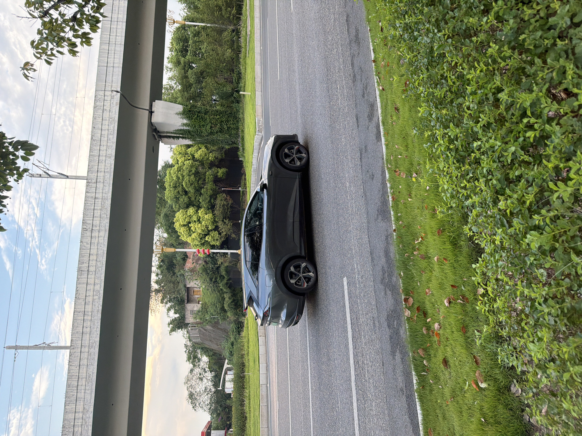 A dark gray car is driving on a paved road in Jiangjin, China. The car is positioned in the right lane and is facing to the left. The road is bordered by a strip of green grass and some bushes. In the background, a large concrete overpass runs horizontally across the frame, supported by concrete pillars. Trees and buildings are visible behind the overpass. The sky is partly cloudy.