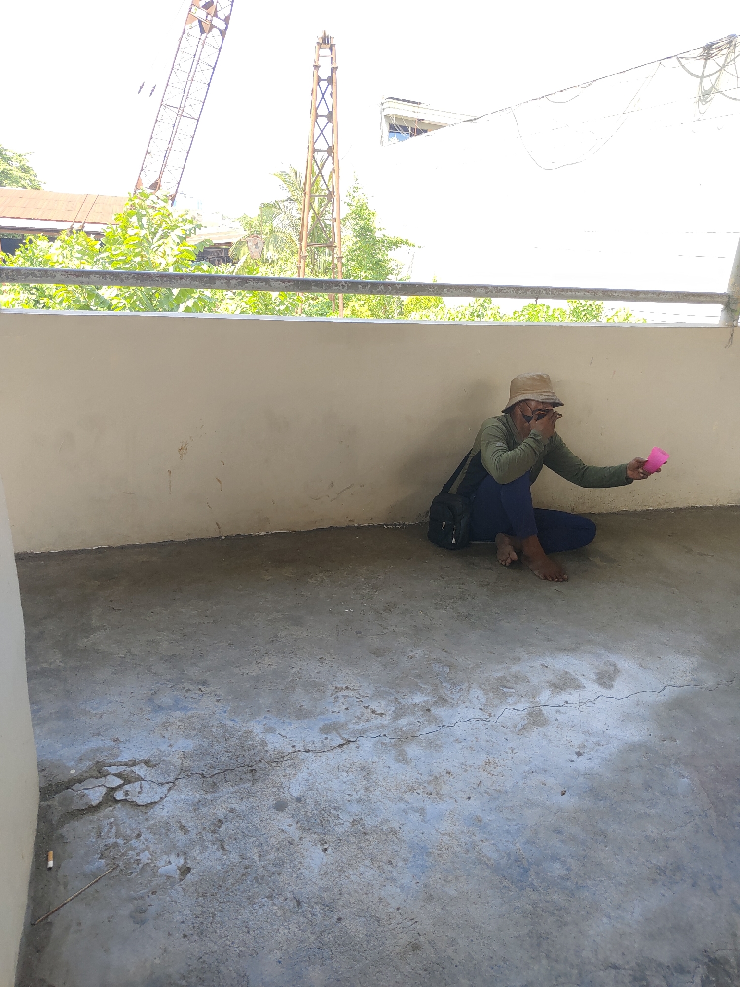 A man wearing a beige bucket hat, sunglasses, a long-sleeved olive green shirt, and dark blue pants is sitting on the concrete floor of a balcony. He is holding a small pink cup in his right hand and appears to be covering his eyes with his left hand. A black shoulder bag is slung across his body. The balcony has a light beige wall behind him and a concrete floor that shows signs of wear and cracking. In the background, beyond a metal railing and green foliage, are industrial structures including a tall crane, suggesting an urban or construction-adjacent environment. The overall lighting suggests it is daytime. The scene is located on a balcony overlooking an area with trees and buildings, consistent with a location in Davao City, Philippines.