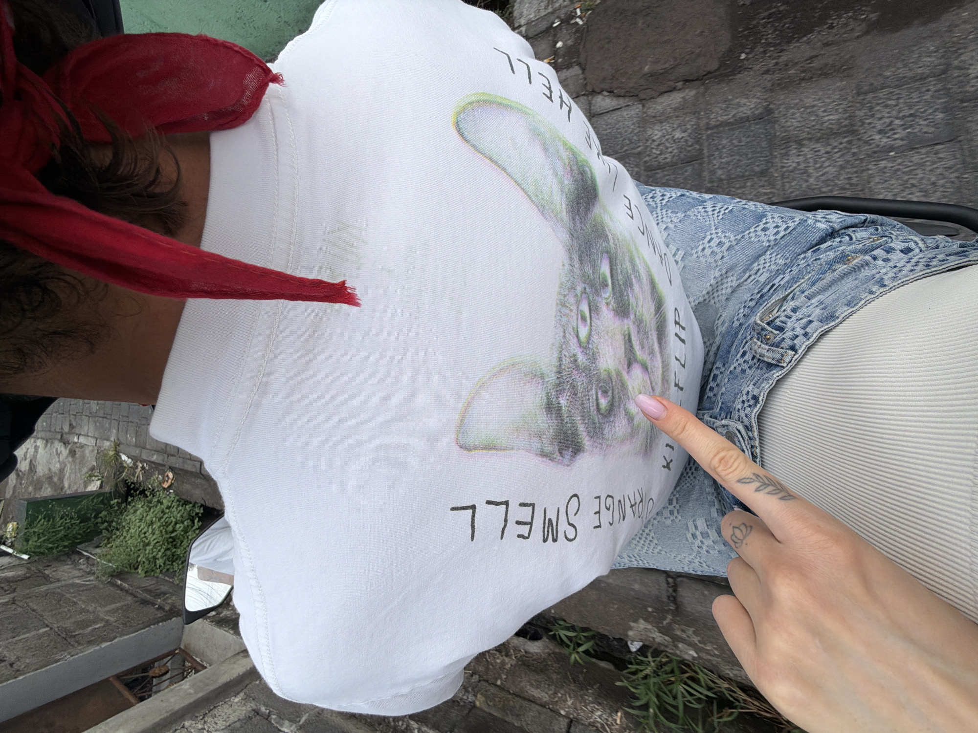 The image shows a person from the back wearing a white t-shirt with a graphic of a cat's face that has a glitch effect. The t-shirt has text printed on it, with "STRANGE SMELL" written vertically on the left side and "HELL" written vertically on the right side. Below the cat's face, there is some more text, partially obscured. The person is wearing blue denim jeans with a patterned texture. A hand with a light pink fingernail is pointing towards the graphic on the t-shirt. There are tattoos on the fingers of the hand, including a lotus flower and a leaf design. The person is seated, possibly on a motorcycle, as indicated by a side mirror visible on the left side of the image, reflecting greenery. The background shows a street or pathway made of gray cobblestones, with a stone wall and lush green vegetation on the left. The location is identified as North Kuta, Indonesia.
