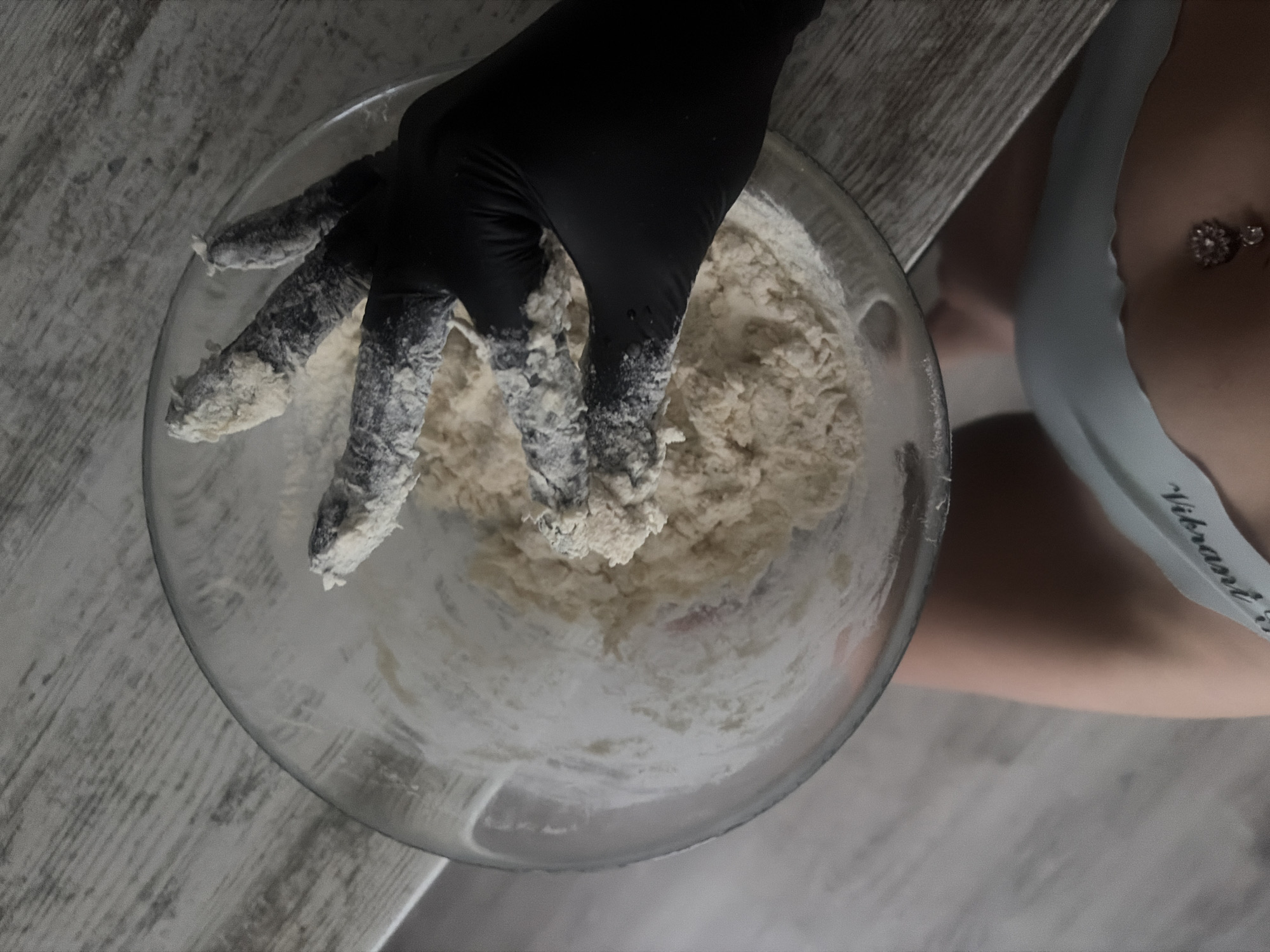 A close-up, overhead shot shows a person's hand, wearing a black glove, reaching into a clear glass bowl filled with dough. The glove and fingers are coated in white flour. The bowl sits on a light-colored, textured surface, possibly a wooden table. Below the bowl, a portion of the person's body is visible, wearing light blue underwear with the word "Vibrant" printed on it. A navel piercing with a floral charm is visible. The background is a blurred, muted color palette. There is no information in the image to indicate a specific location in the city of Unknown, Russia.