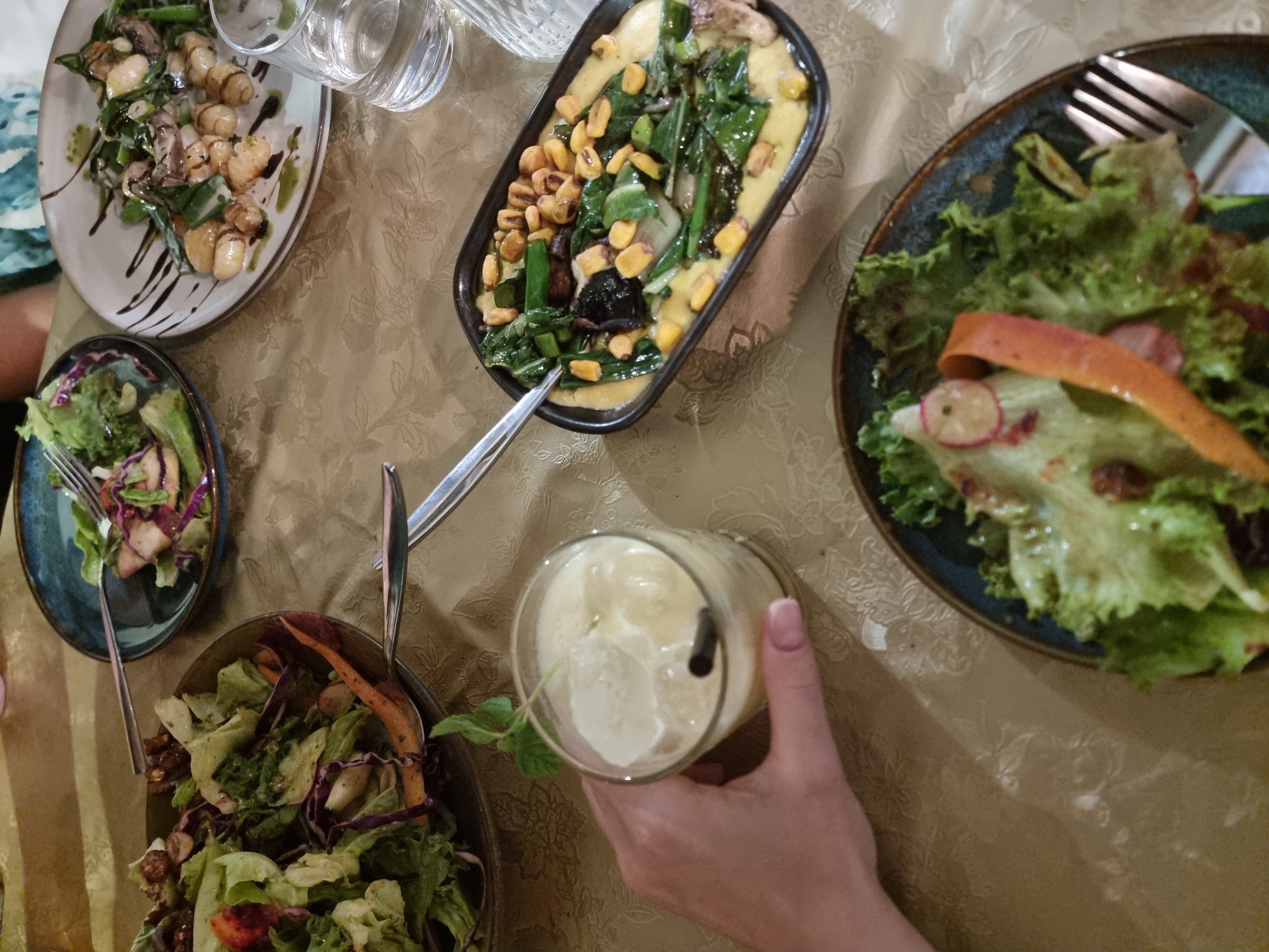 A table laden with various food dishes is seen from an overhead perspective. Dishes include salads, gnocchi with dark sauce drizzle, a corn and greens dish with a creamy yellow base, and another salad on a smaller plate. Glassware, including a water carafe and glasses, are present. A hand is holding a glass with a light-colored beverage, ice, and a straw. The table is covered with a patterned, light-colored cloth. The location is suggested as Mitzpe Ramon, Israel.