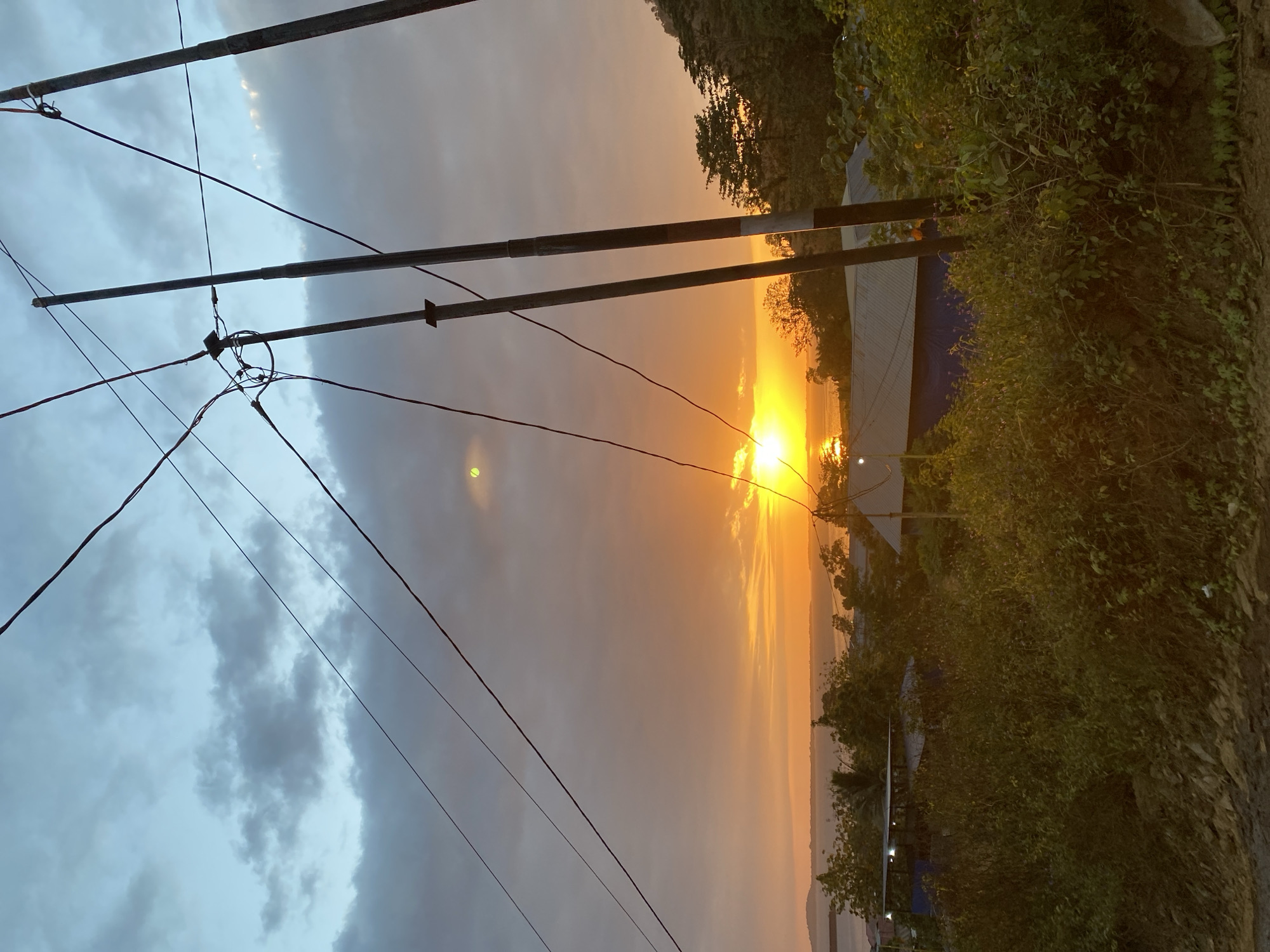 The image depicts a sunset scene from a slightly elevated perspective in Pulau Bapa, Indonesia. The sky is a gradient of warm oranges and yellows near the horizon, transitioning to cooler blues and grays with scattered clouds overhead. A bright sun is visible just above the horizon, casting a strong reflection on the water. In the foreground, a tangle of green bushes and vegetation covers a rocky slope. Power lines, supported by wooden poles, stretch across the upper portion of the image. In the middle ground, several buildings with corrugated metal roofs and some with blue tarpaulin coverings are nestled among trees. The water body, likely a bay or strait, is visible in the distance, with a faint outline of land on the horizon. The overall atmosphere is tranquil, capturing the end of the day. No people are visible in the frame.
