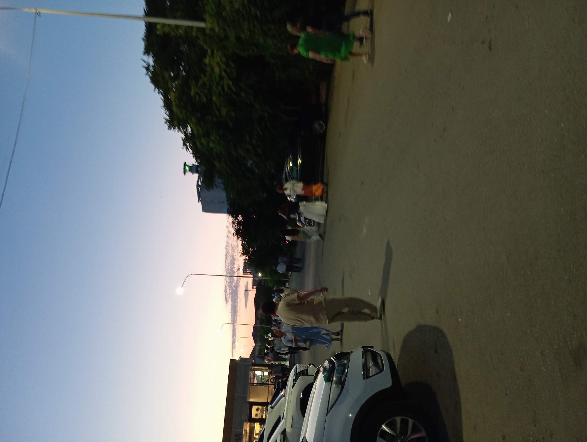 A street scene in Aktau, Kazakhstan, shows a group of people walking along a paved road during dusk. The sky is a pale blue, transitioning to a soft pink and orange near the horizon, indicating the sunset. Streetlights are visible, and one is illuminated, casting a glow. On the left side of the image, several cars are parked. A white SUV is prominently in the foreground, with its front end visible. Further back, more cars are parked near a building with a modern facade and large windows. The people in the image are blurred due to motion, but appear to be casually dressed. Some are walking individually, while others are in small groups. A man in a light-colored shirt and trousers is walking away from the camera. To his left, a woman in a light blue dress is walking. Further down the road, there are more figures and parked cars. Trees line the right side of the road, with dense foliage. In the background, a tall building with a green light on top is partially visible. The overall atmosphere is relaxed, suggesting a public gathering or evening stroll.