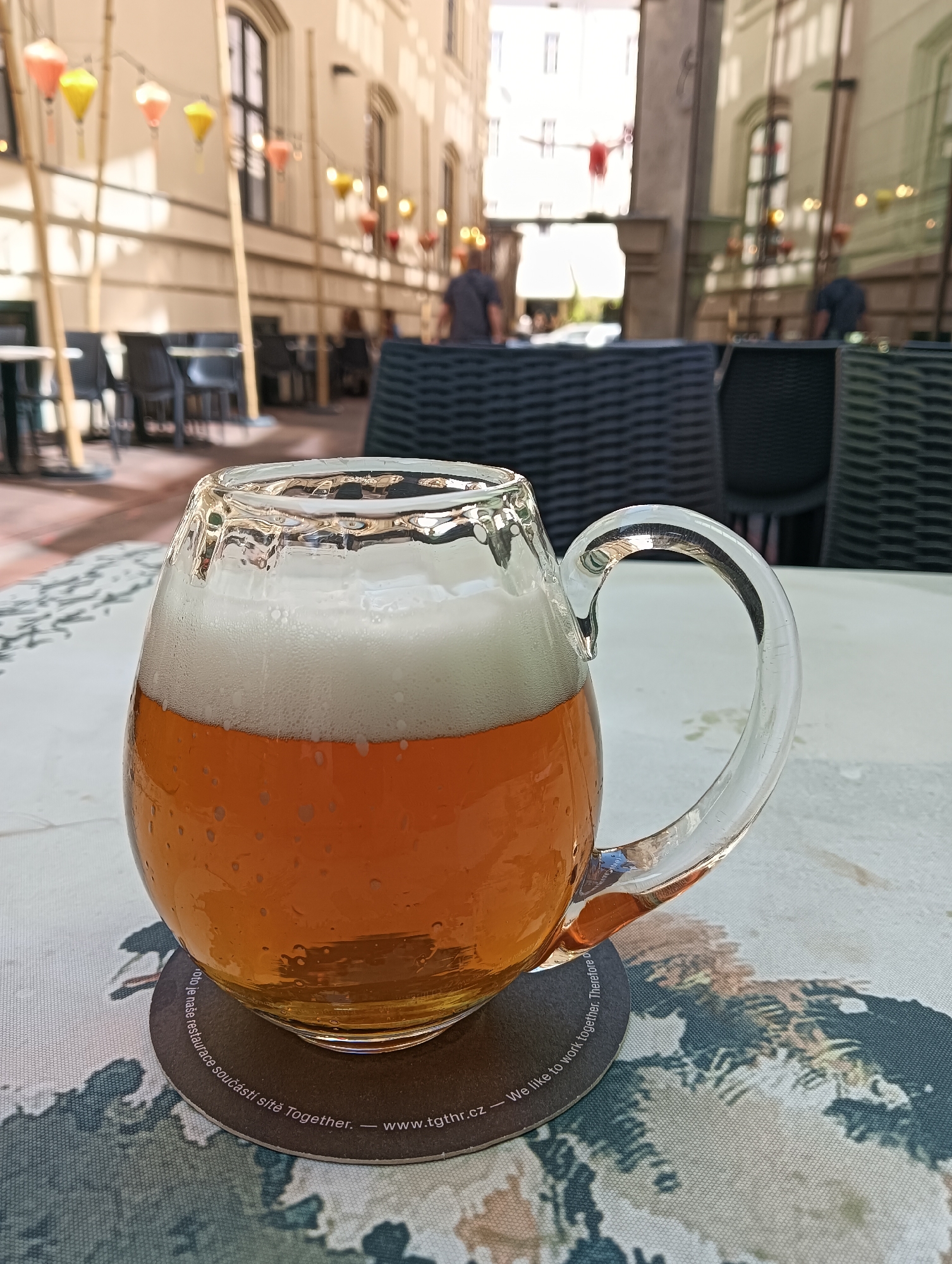 A glass mug of amber beer with a thick white head of foam sits on a table with a patterned tablecloth. The mug is on a dark coaster with white text that reads "Jsme rádí, že jste součástí sítě TOGETHER. - www.tgthr.cz - We like to work together. Therefore we...". In the background, an outdoor seating area of a restaurant is visible. Tables and chairs are set up under awnings, with strings of decorative lanterns overhead. Several people are blurred in the background, and architectural elements of buildings line the street. The scene appears to be in an urban setting, likely a restaurant or cafe in Prague, Czechia, given the text on the coaster.