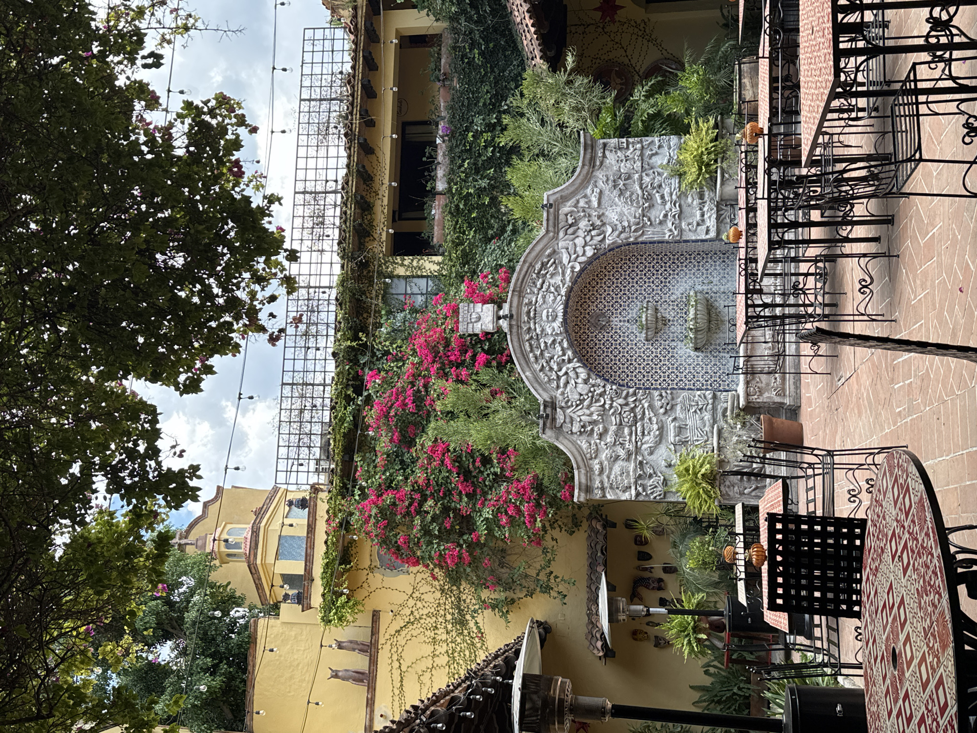 The image captures a scene in San Miguel de Allende, Mexico, in an outdoor courtyard setting. A stone fountain with intricate carvings and blue tile accents serves as a focal point. Red bougainvillea vines drape down the adjacent wall of a yellow building. Black wrought iron tables and chairs are positioned on a brick patio. Green trees and foliage are present, adding a natural element to the urban environment. Decorative items adorn the walls, contributing to the cultural and artistic ambiance. Overhead lights are strung across the courtyard.