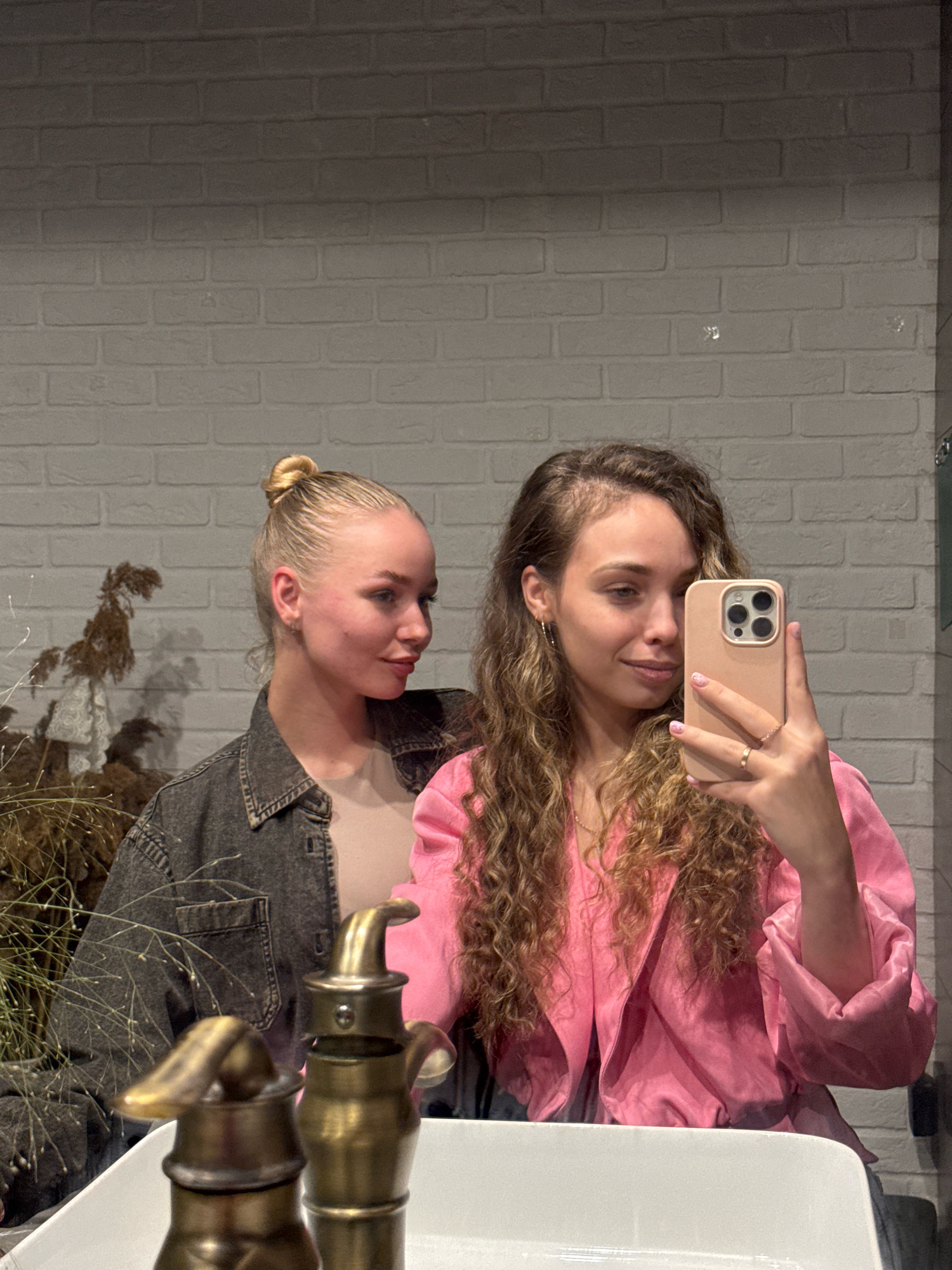 Two young women are taking a selfie in a bathroom with white brick walls. The woman on the right is holding a pink smartphone and wearing a pink satin jacket. The woman on the left has her hair in a bun and is wearing a denim jacket. They are both looking into the phone's camera. A gold-colored faucet is visible in the foreground, along with some dried reeds and branches in the background. The image appears to be taken in Chaikovsky, Russia.