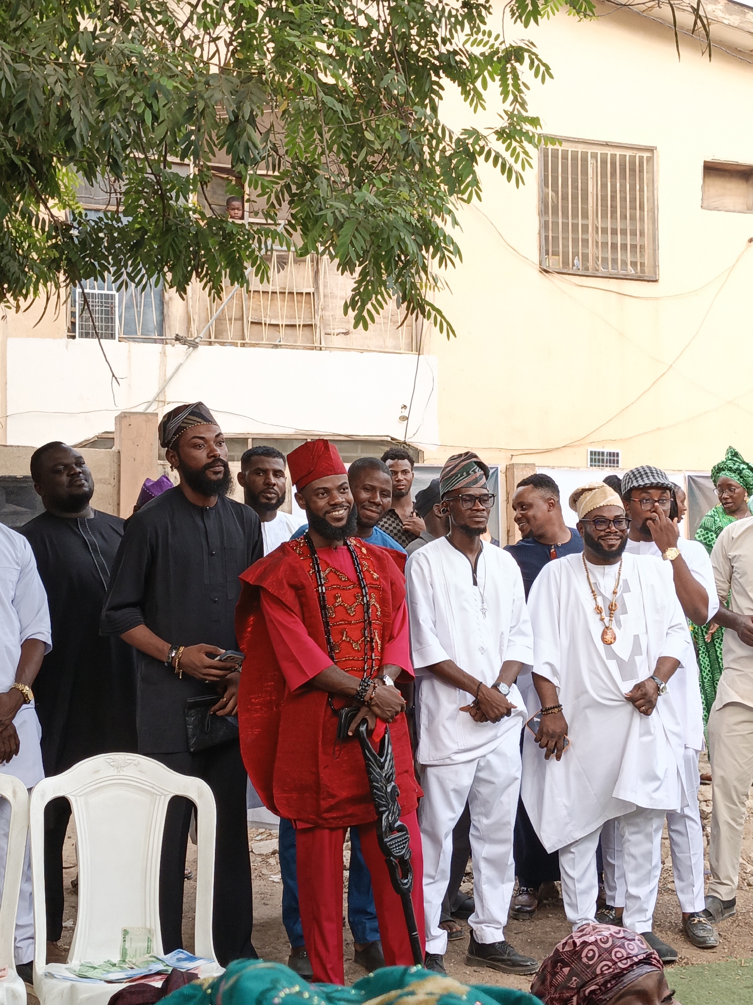 An outdoor gathering in Gafan, Nigeria, depicts a group of men dressed in traditional formal attire, suggesting a cultural celebration or social event. The majority of the approximately fifteen men are young to middle-aged, standing in an open, unpaved area.
In the foreground, a prominent man is distinguished by his vibrant red long-sleeved tunic and trousers, adorned with multiple layered beaded necklaces in red, black, and white, and a matching red cap. He holds an ornate, dark staff or cane. Many other men wear flowing white tunics and trousers, complemented by various patterned caps (fila) and some wear glasses. One man wears a black tunic and trousers, also with a patterned cap, holding a mobile phone. Another individual, partially visible on the far right, wears a green headwrap and attire. Most subjects exhibit positive emotions, with smiles and attentive expressions, generally looking towards the left side of the frame.
Objects in the scene include white plastic chairs; on one, several currency notes (appearing to be Nigerian Naira) are neatly stacked. The background features a light-colored building with barred windows, partially obscured by lush green leafy trees. A child's head is visible peeking out from one of the upper windows through the foliage. The lighting suggests it is late afternoon or early evening, casting a warm glow. No discernible text is visible in the image.