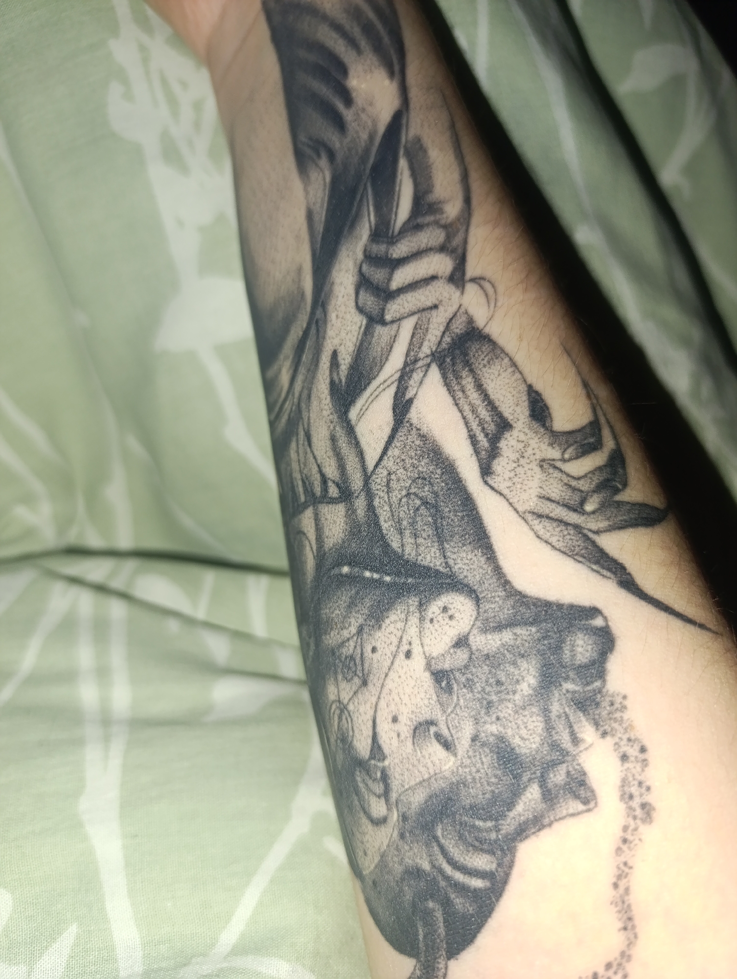 A close-up image shows a forearm tattoo. The tattoo is black and white and depicts a figure holding a scythe. The forearm is resting on a light green fabric with a faint floral pattern. The scene is located in a closed environment.