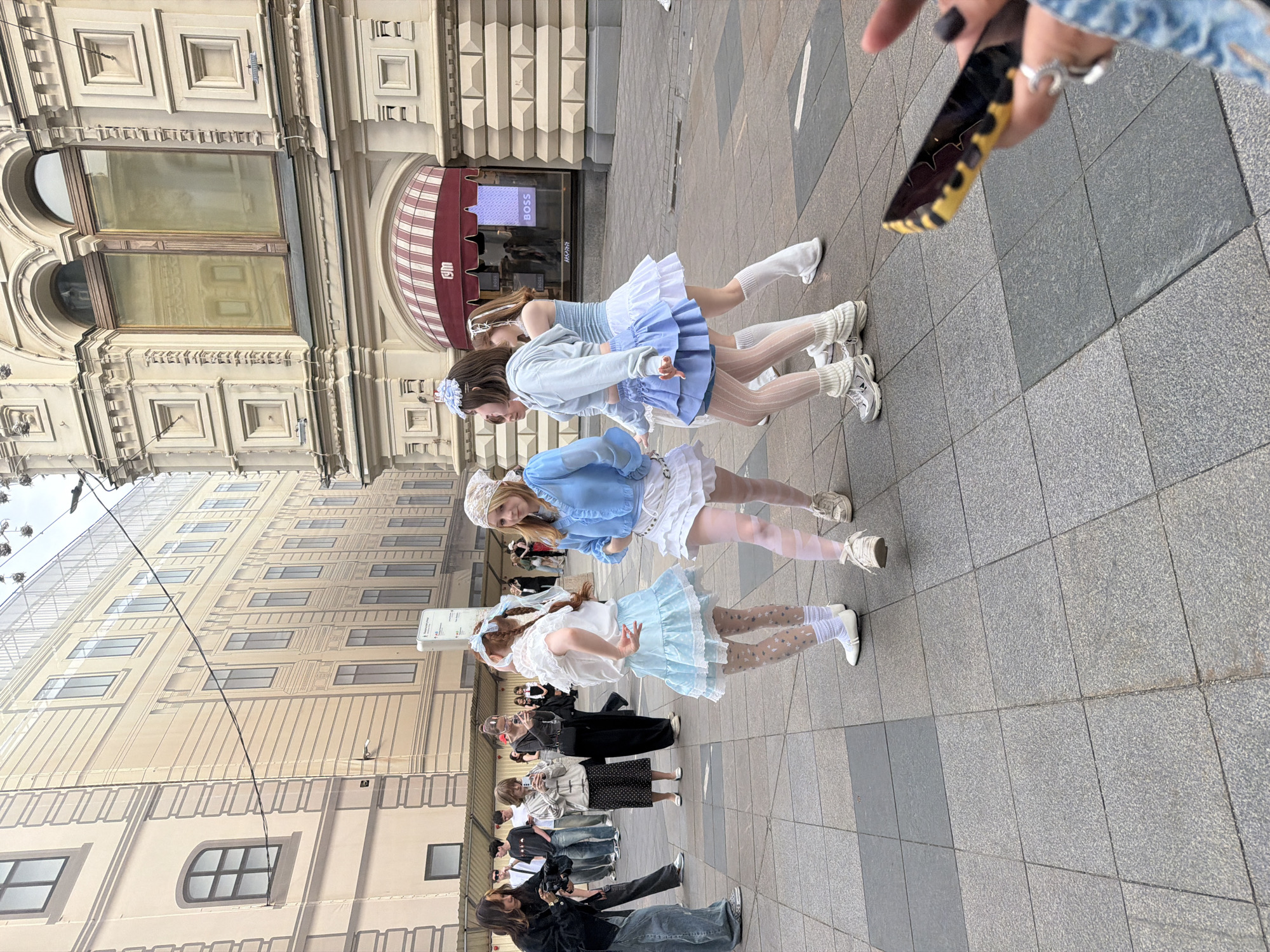 The image shows four young women posing in front of the GUM department store in Moscow, Russia. They are dressed in light-colored, frilly dresses with blue accents and white socks and sneakers. A crowd of people, some holding cameras, are watching and photographing them. The GUM building is visible in the background with its red and white striped awnings. The ground is paved with grey tiles. Someone is holding up a cellphone to possibly take a photo or record a video.