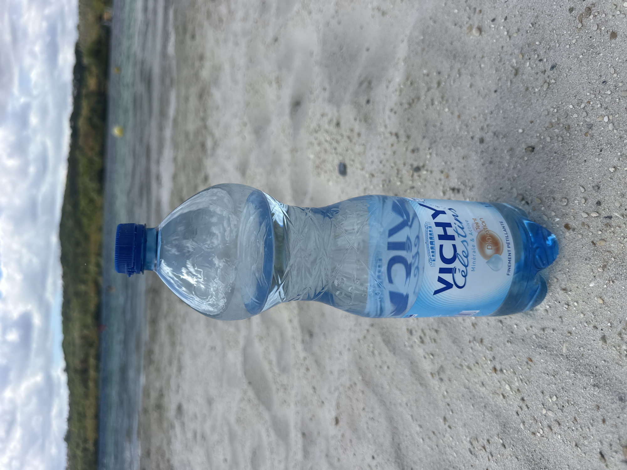 A clear plastic bottle of Vichy Célestins mineral water with a blue cap stands on a sandy beach. The bottle is half-full and has a blue and white label with French text. In the background, a body of water is visible, with a green, vegetated hill on the horizon. The sky is cloudy. The scene appears to be a natural outdoor setting, possibly a lake or the sea. The location is specified as Jablines, France.