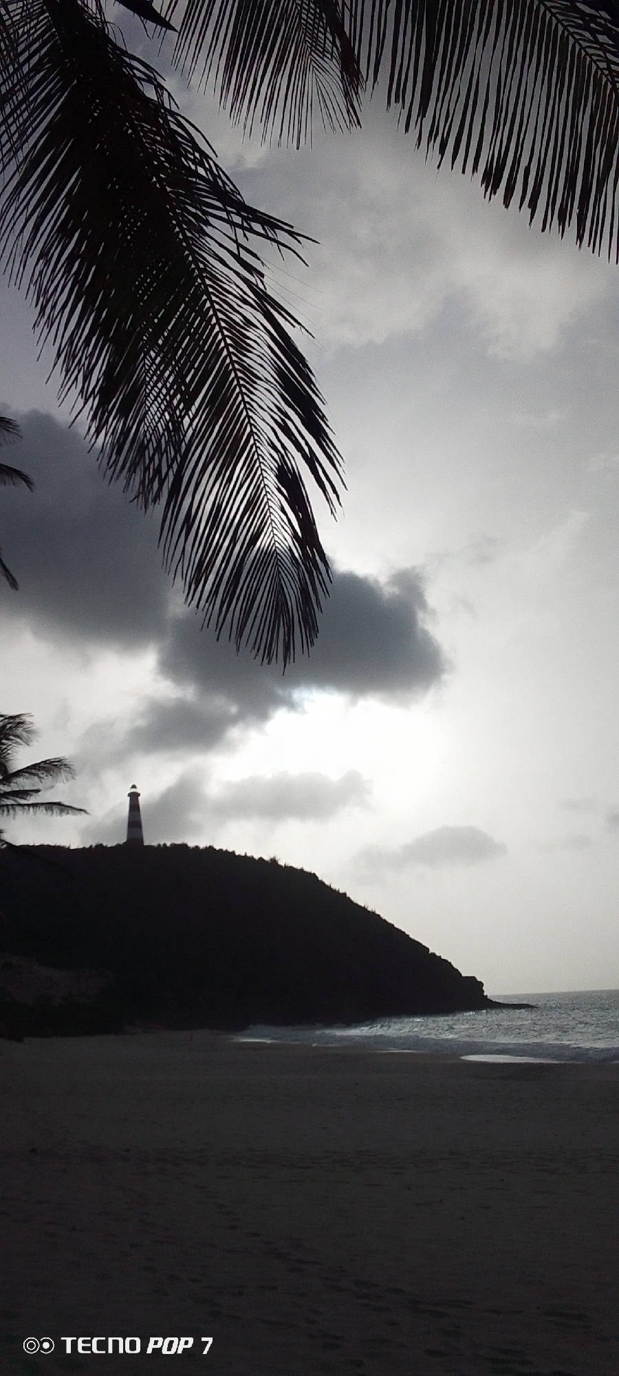 The image shows a grayscale view of Cabo de la Caracola, Pedro González, Venezuela. Palm tree fronds overhang the top of the frame. In the distance, a lighthouse sits atop a dark, silhouetted hill. The sky is overcast with a bright spot where the sun breaks through the clouds. A sandy beach stretches across the lower portion of the frame, meeting the ocean waves.