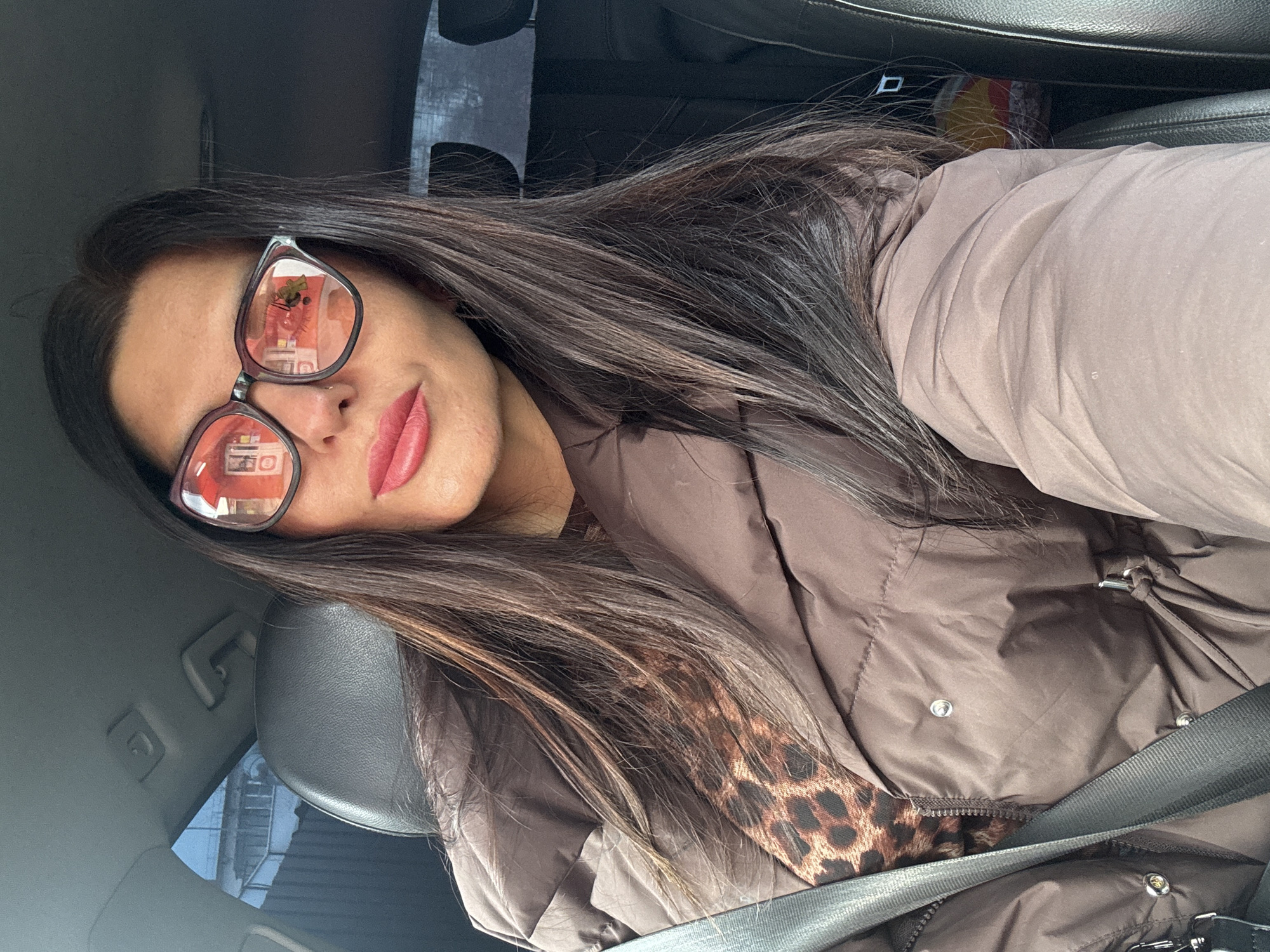 A woman wearing glasses and a brown jacket with a leopard print collar is sitting in a car, fastened with a seatbelt. The glasses reflect buildings and signage in the city of Chaikovsky, Russia.