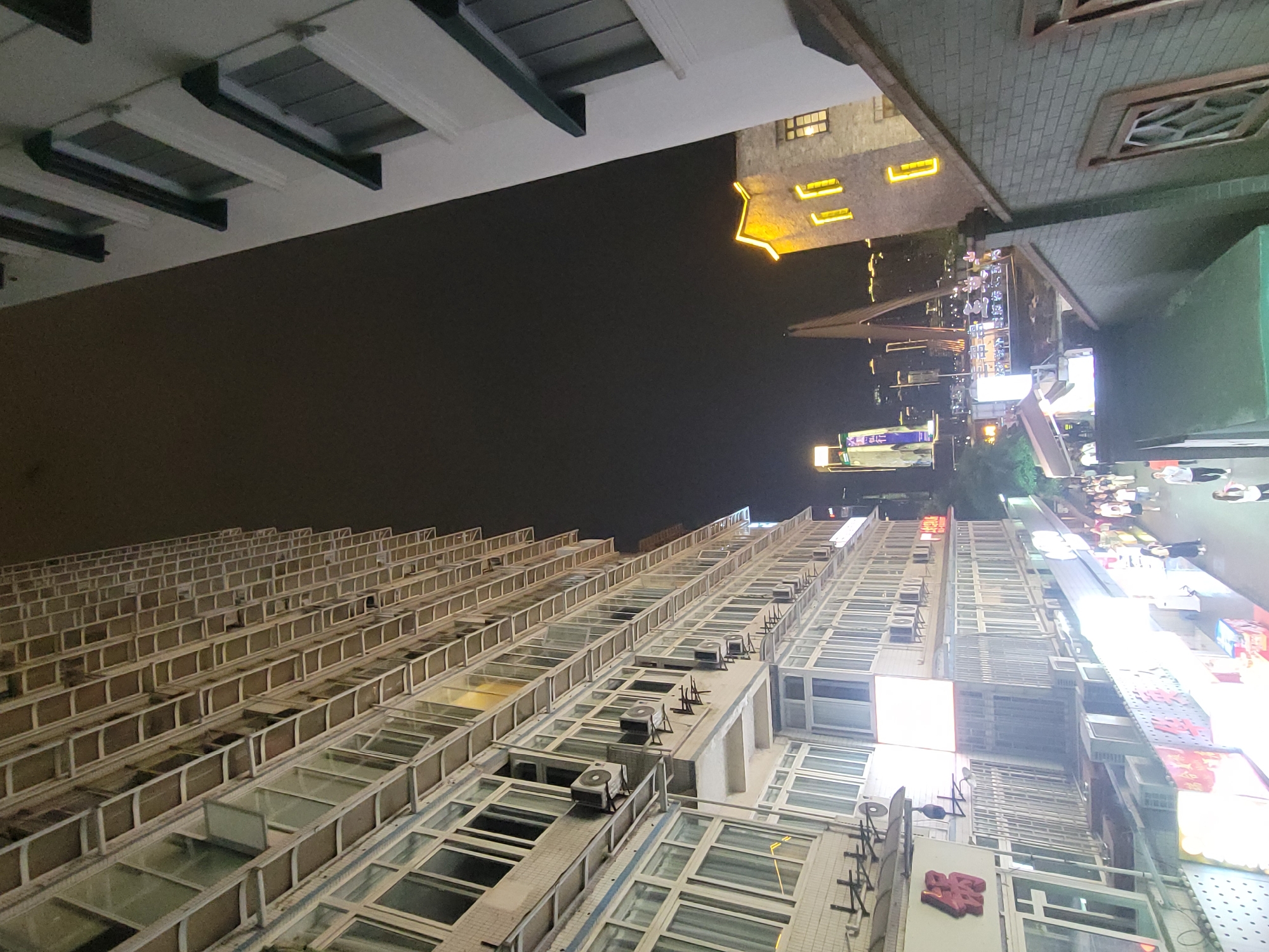 This night-time photograph captures an upward-looking view of a dense urban environment, likely in Yuzhong District, China, as specified. The image is oriented with the ground level on the right side and the dark sky on the left.
On the left, a tall, modern high-rise building dominates, characterized by numerous uniform windows with light-colored frames, many featuring small balconies or ledges. Several outdoor air conditioning units are visible along its facade.
To the right, the architecture shifts to more ornate structures. One building features a dark, brick-patterned facade with a decorative, possibly traditional, window. Below and further right, another building stands out with a distinctive yellow-lit roofline and several bright yellow rectangular lights. These structures suggest commercial or entertainment venues.
The far right and bottom of the frame reveal a busy street scene, brightly illuminated by various neon and LED signs. Numerous indistinct figures of people are visible on the street level, appearing to walk and congregate, indicative of active night life. Blurred Chinese characters are present on several illuminated signs; a prominent red sign partially displays the character "翠" (cuì). The dark, clear night sky provides a stark contrast to the intense artificial lighting from the buildings and street below, highlighting the bustling urban landscape.