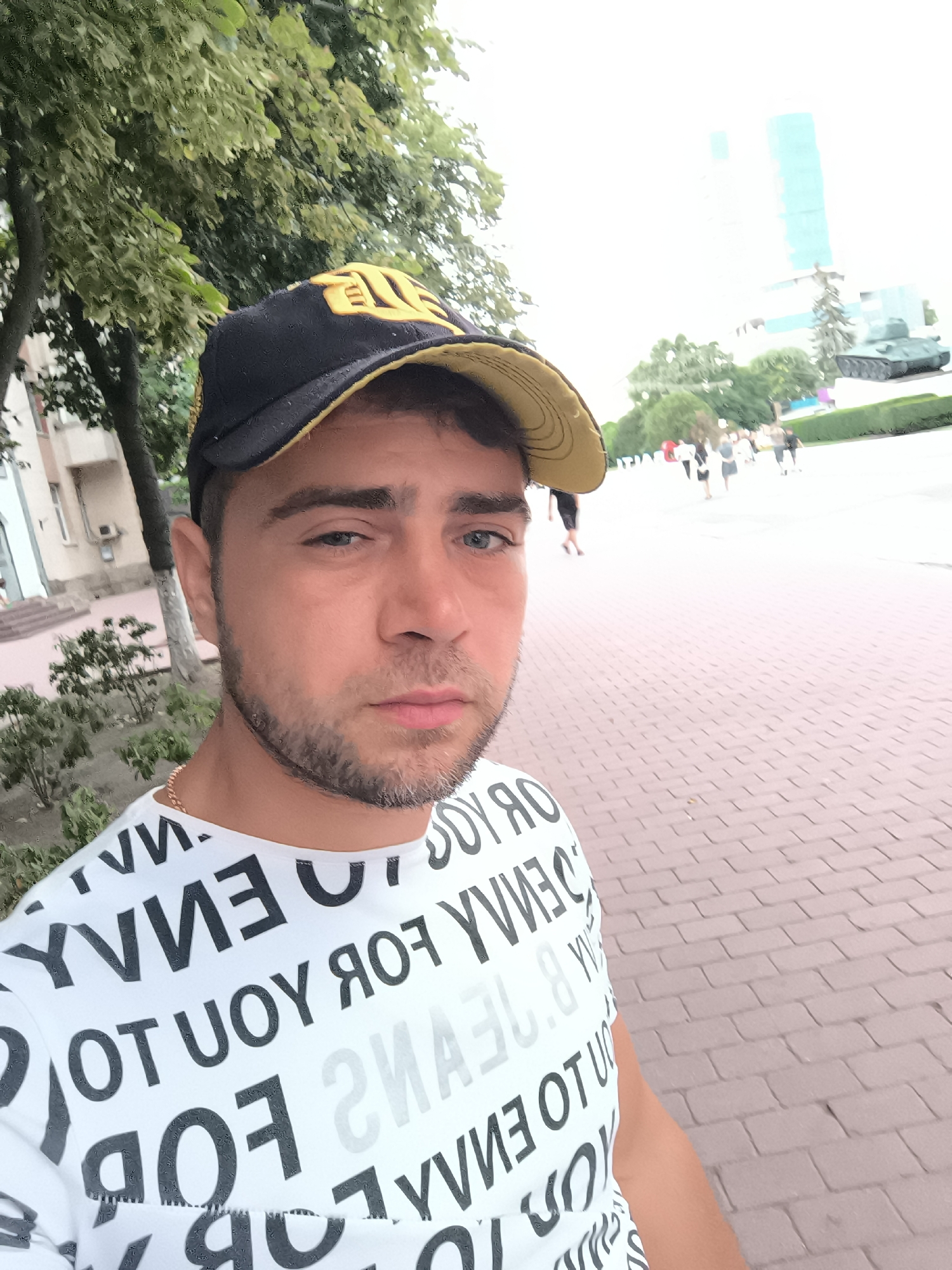 A medium shot shows a man in a black and yellow baseball cap and a white t-shirt with text. The man is facing the camera, and his left eye is visible. He has a beard and short hair. He is wearing a gold chain. In the background, there are trees, buildings, a tank statue, and people walking on a brick sidewalk. The lighting is bright, suggesting daytime.