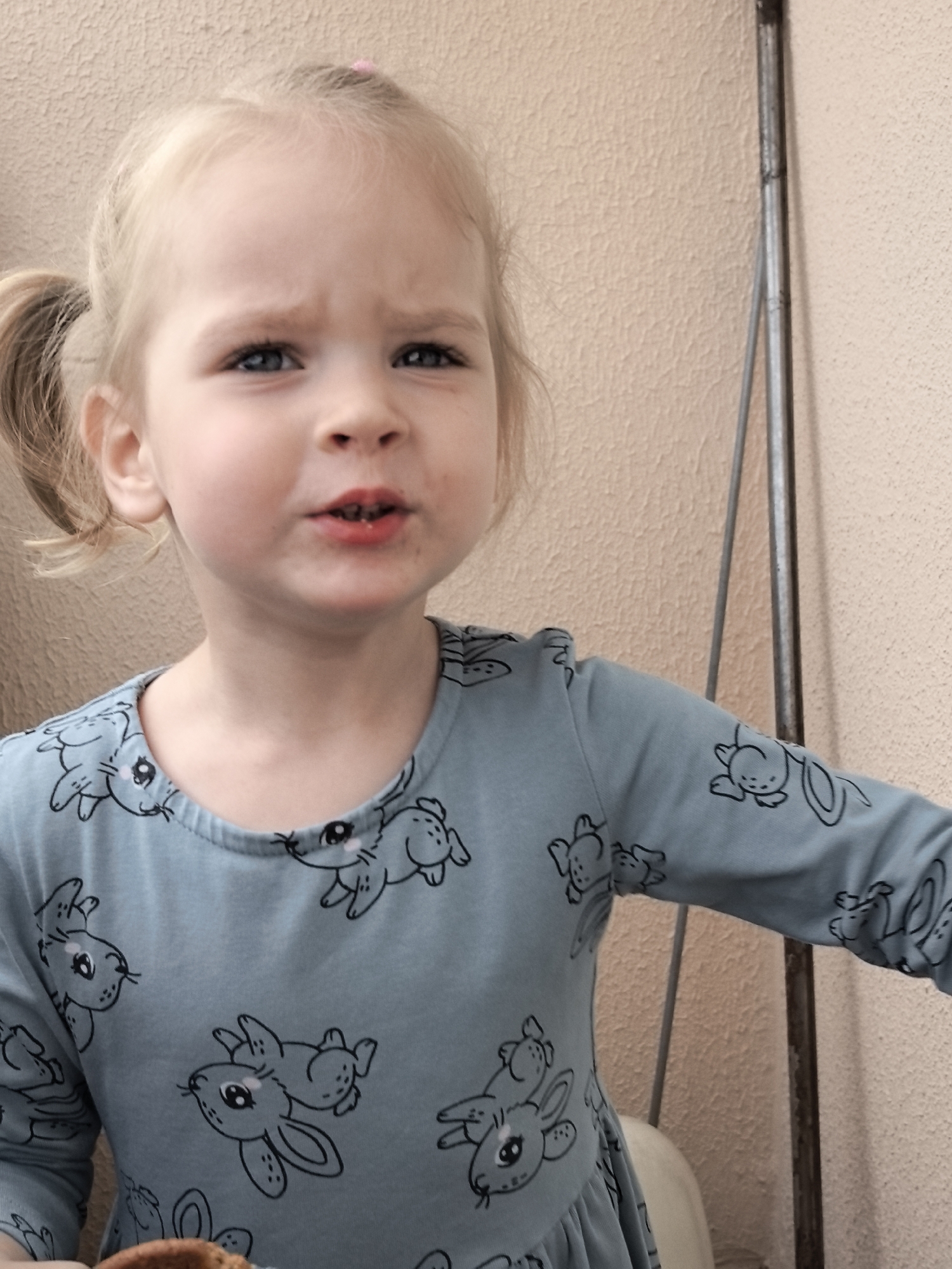 A young Caucasian girl with blonde pigtails is looking towards the camera with a displeased expression. She is wearing a light blue long-sleeved shirt adorned with a pattern of black bunnies. Her mouth is slightly open, revealing some food particles around her lips. The background consists of a textured, light peach-colored wall and a narrow, vertical, metallic object on the right side of the frame. The lighting appears to be natural daylight, casting soft shadows. The image was taken outdoors in an unknown city and country.