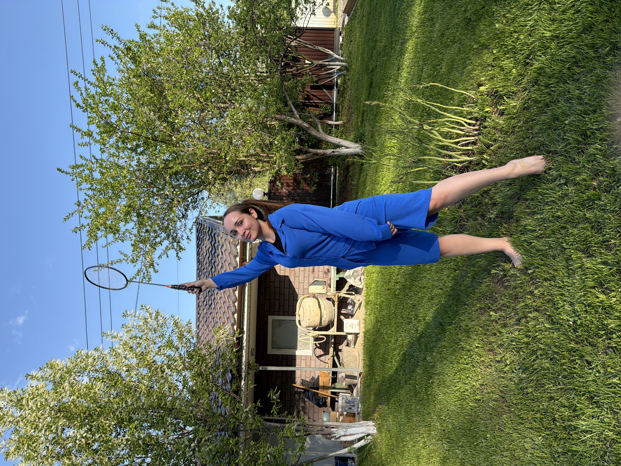 A woman lies horizontally on a green lawn in what appears to be a backyard in the city of Chaikovsky, Russia. She is dressed in a blue outfit. She holds a badminton racket in her extended right hand. Her feet are bare. The background includes a tree, a house with a brown roof, a power line, garden plants, and various outdoor objects, possibly construction materials or tools. The sky is blue.