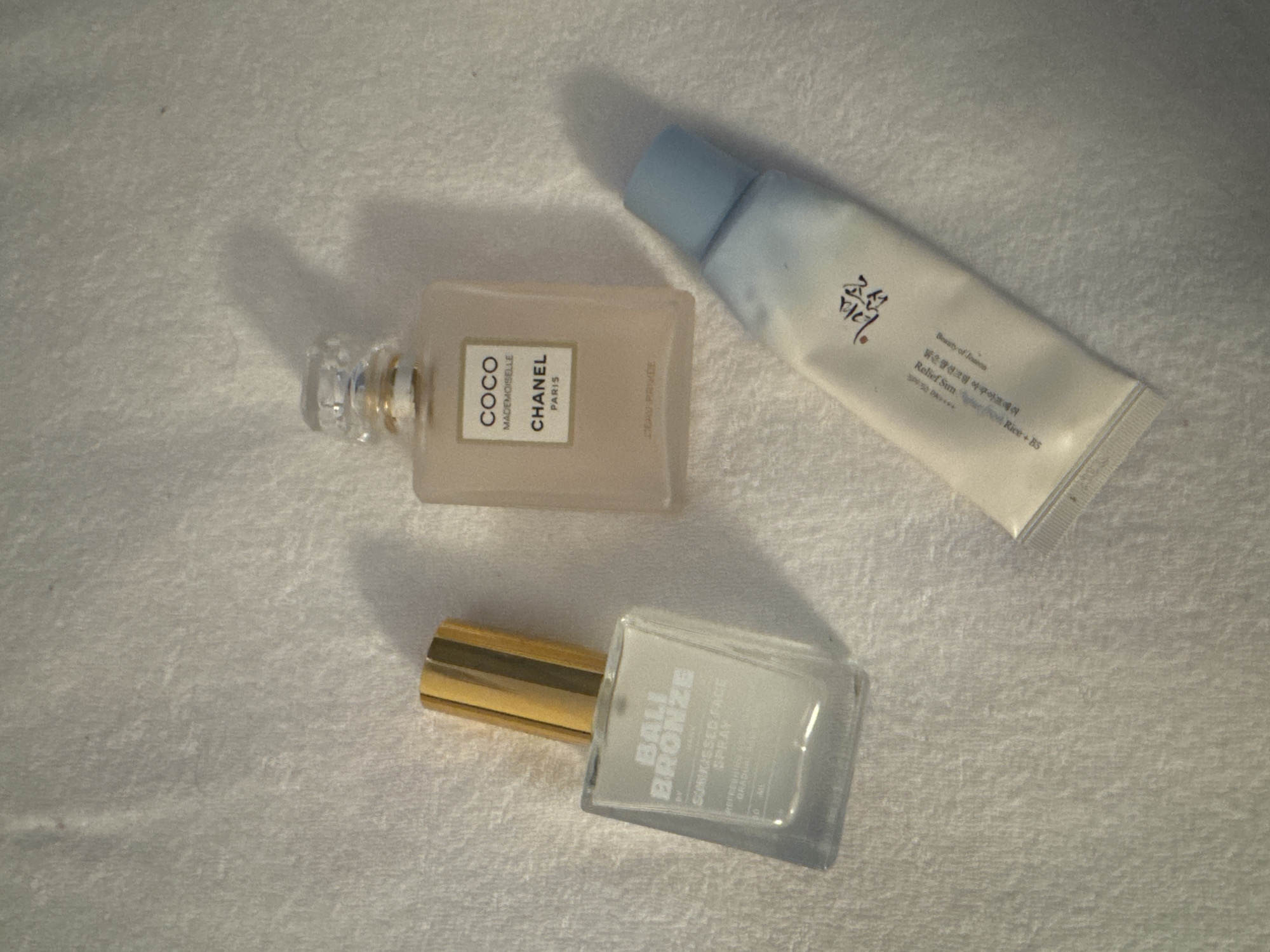 The image shows three cosmetic products laid out on a white textured surface. From left to right, the first object is a clear glass spray bottle with a gold cap, labeled "BALI BRONZE Sunkissed Face Spray". Next to it is a rectangular pink glass perfume bottle with a clear cap, labeled "COCO MADEMOISELLE CHANEL PARIS". The third object, at the bottom right, is a white squeeze tube with a light blue cap, featuring Korean text and English labels that are partially visible, indicating "Relief Sun" and "Beauty of Joseon". The lighting casts shadows from the objects onto the surface. The location is not specified in the image or provided context.