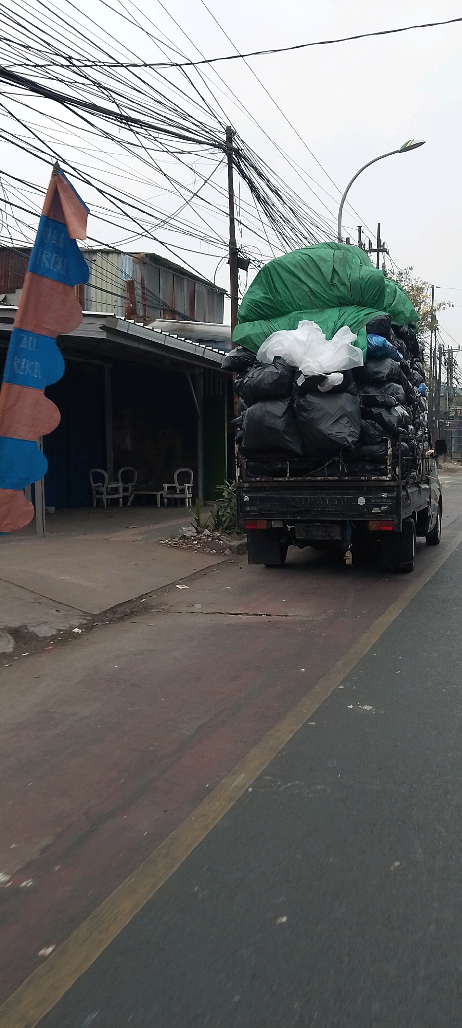 The image captures a street scene in Bekasi, Indonesia, where a pickup truck is heavily laden with black trash bags and topped with a large green tarp and some white plastic. The truck is in motion on a paved road with a yellow line marking the lane. To the left of the truck, there's a structure with a corrugated metal roof and an open-fronted area with two white chairs and a table. A large, vertically oriented banner with alternating orange and blue sections is also visible on the left side of the frame. Overhead, a dense network of power lines crisscrosses the sky, supported by poles, and a street light is mounted on one of these poles. The sky is overcast. The overall environment appears to be an urban or semi-urban area, likely in a developing region given the power line infrastructure and the type of truck and building. The scene is devoid of people.