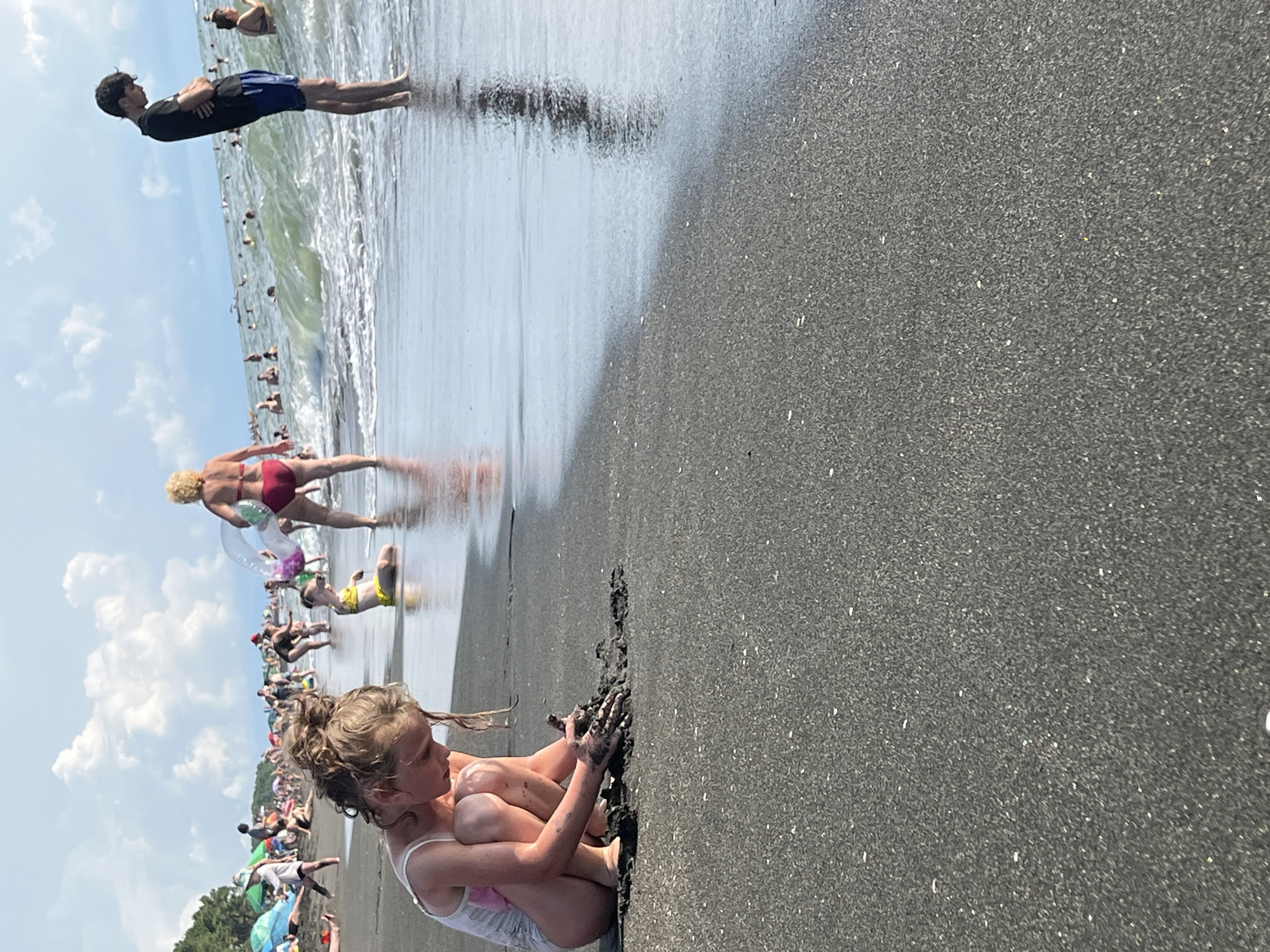 This is a high-angle, full-shot photograph taken at eye-level on a beach. The image is rotated 90 degrees counter-clockwise. A young girl with blonde hair is in the foreground, squatting and playing with black sand. She is wearing a white swimsuit. In the middle ground, a woman with blonde hair in a red bikini is walking into the water, holding a clear inflatable ring. Further in the background, several people are in the shallow water, some swimming and others standing. The sky is blue with scattered white clouds. The beach appears to be a mix of dark sand and wet sand. The overall scene depicts a sunny day at the beach with people enjoying the water and sand. The location appears to be Ureki, Georgia, known for its magnetic black sand beaches. There is no visible text in the image.