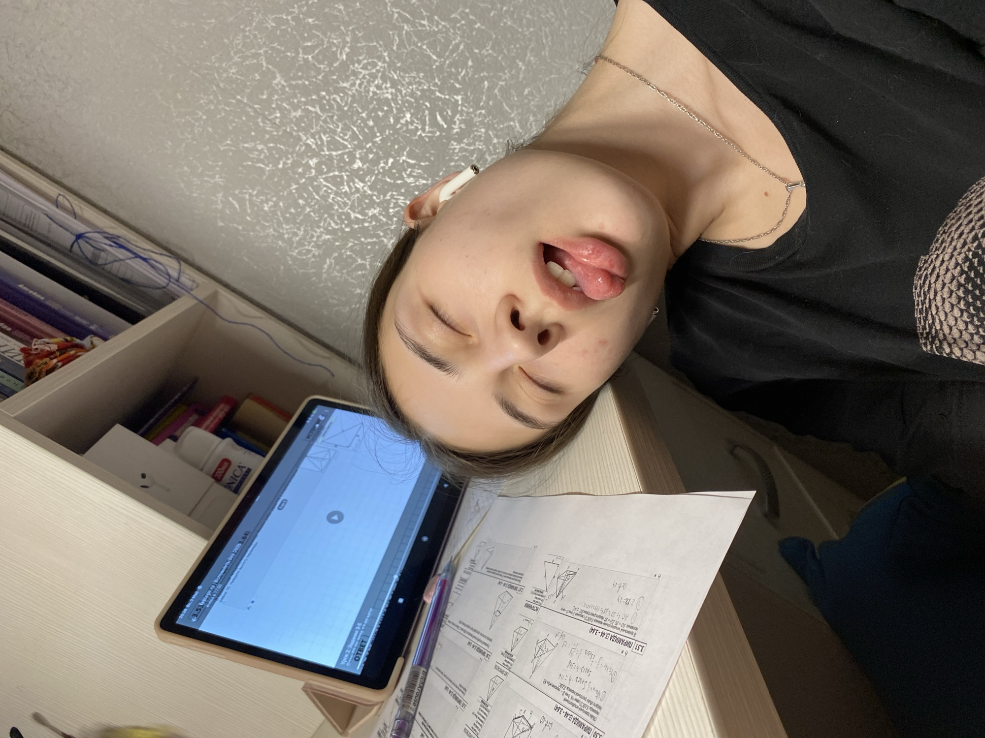 A young woman is lying on a desk with her eyes closed and her tongue sticking out. She has an earbud in her left ear and is wearing a black shirt and a silver necklace. On the desk are a tablet displaying geometric drawings, papers with geometric problems, and a pen. A shelf to the left of the desk contains books and other items, including a white box and a bottle of lotion. The background is a textured light gray wall. The location in the city of Votkinsk, Russia, is not discernible from the image.
