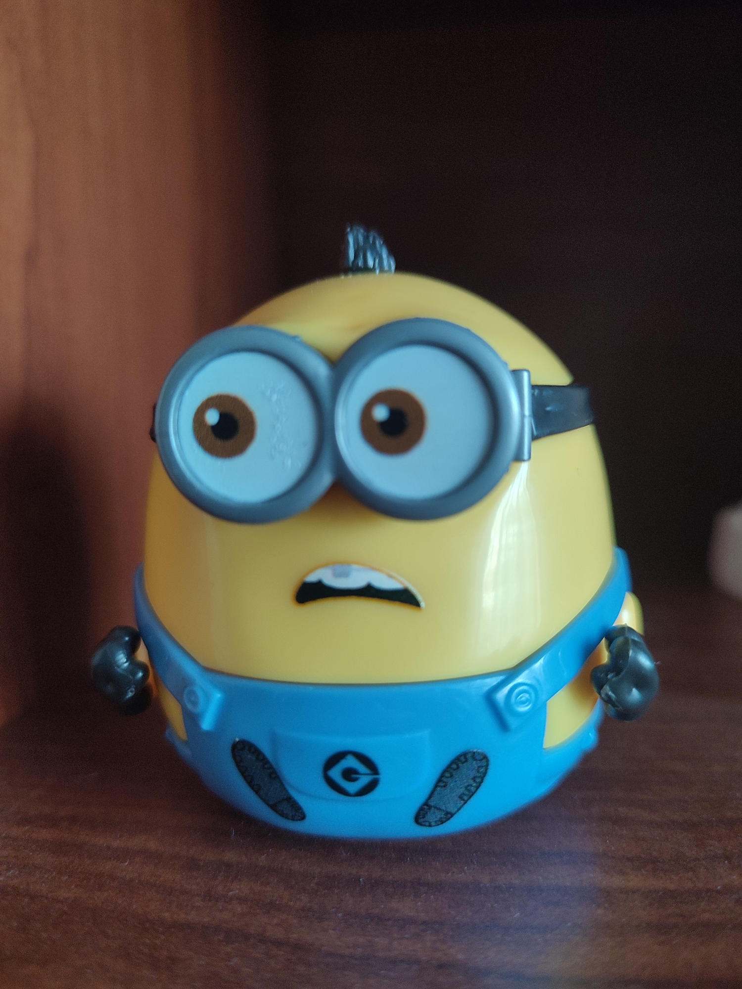 A close-up, eye-level, medium shot captures a yellow Minion toy with a blue jumpsuit and goggles. The toy is positioned on a wooden surface, likely a shelf or table, with a blurred brown background. The toy's mouth is open in a distressed expression, and its eyes are wide open. The lighting is natural, creating soft shadows. There is no indication of the city of Voronezh, Russia, or any other geographical location.