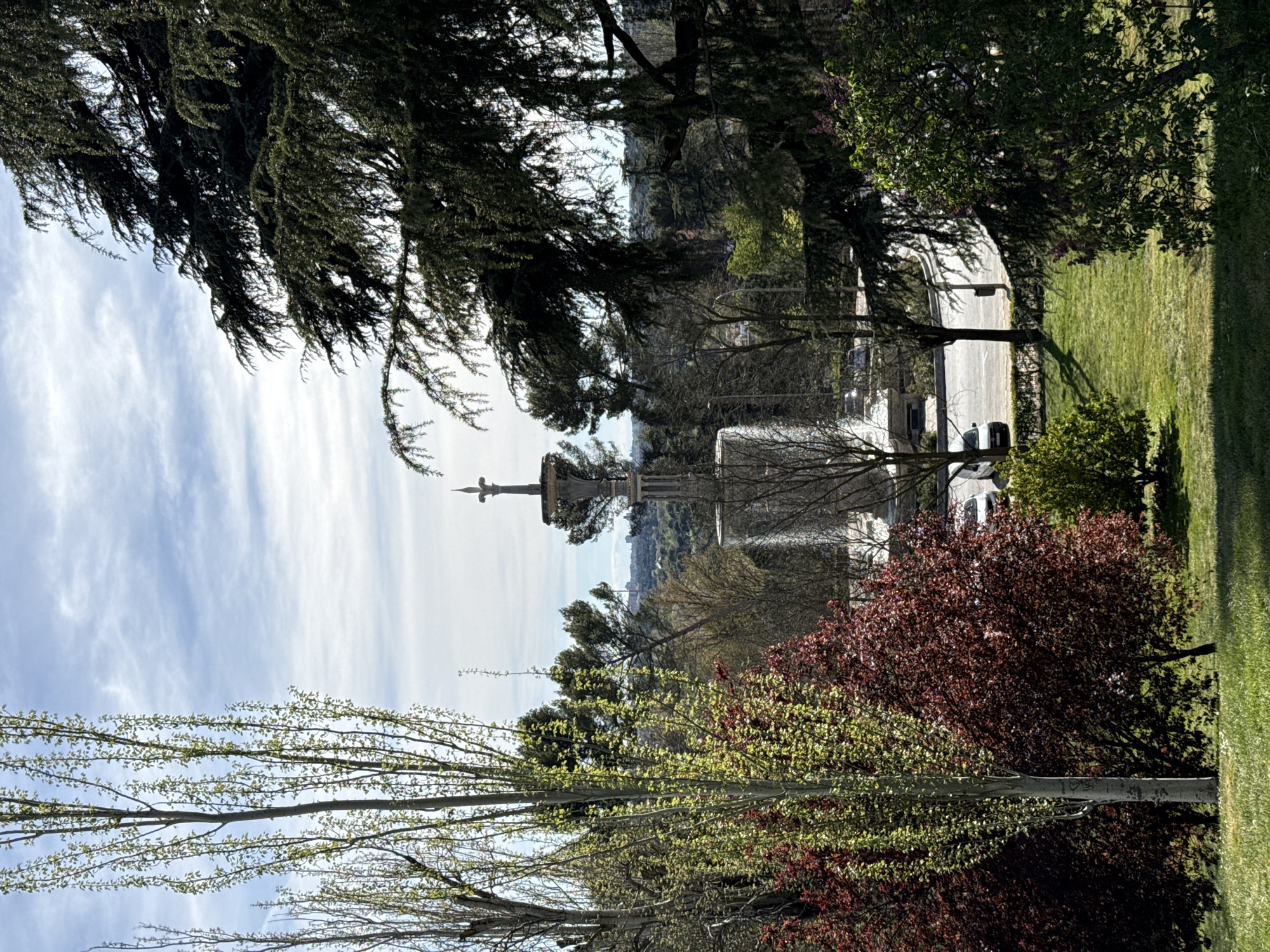 The image shows a view in Madrid, Spain, looking towards the Monument to Cristobal Colon at the Plaza de Colón. The monument is visible through a frame of trees, with a mix of deciduous and evergreen varieties. The sky is partly cloudy. A street runs parallel to the viewer, with cars parked along it. In the foreground, trees with budding leaves, and red and green bushes are visible. The time of year is likely spring. The image is shot from a slightly elevated position.