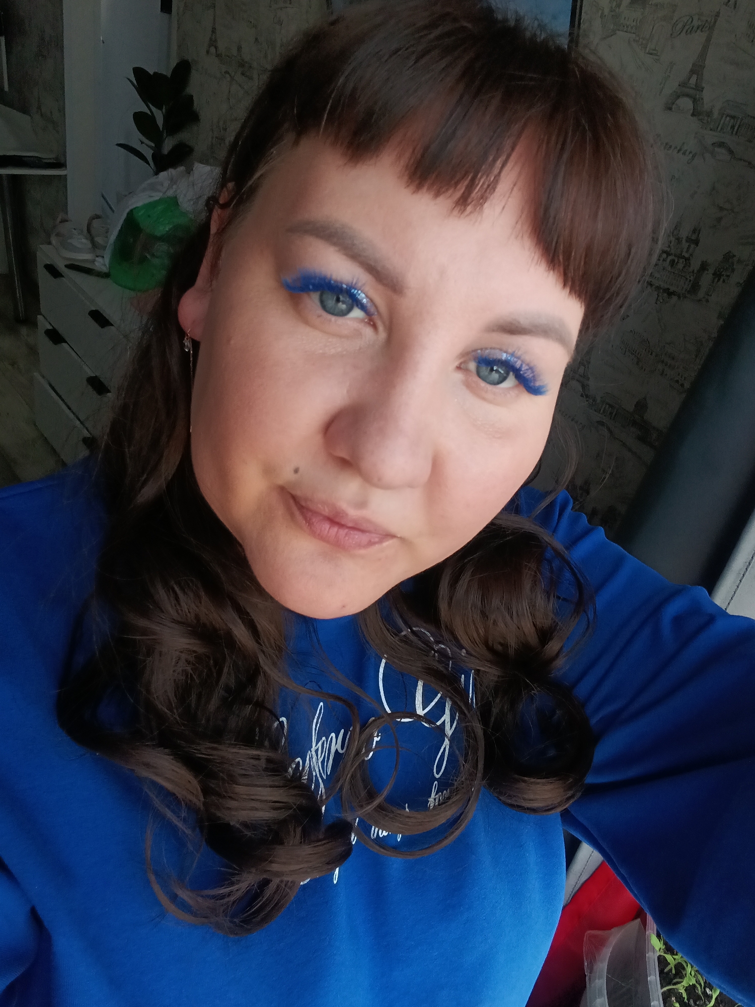 A woman with shoulder-length brown hair with blunt bangs, wearing a blue long-sleeved sweatshirt with white lettering, is captured in a selfie-style, close-up image. The woman has fair skin, blue eyes accentuated with bright blue mascara, and a mole on her left cheek. The background includes a wall with a gray wallpaper featuring an Eiffel Tower pattern. A partially visible white dresser stands against the wall on the left, with a plant on top of it. At the bottom right, there's a blurred view of what seems to be a container with small green plants. While the woman appears to be indoors, the exact location within Osa, Russia is indiscernible from the image.