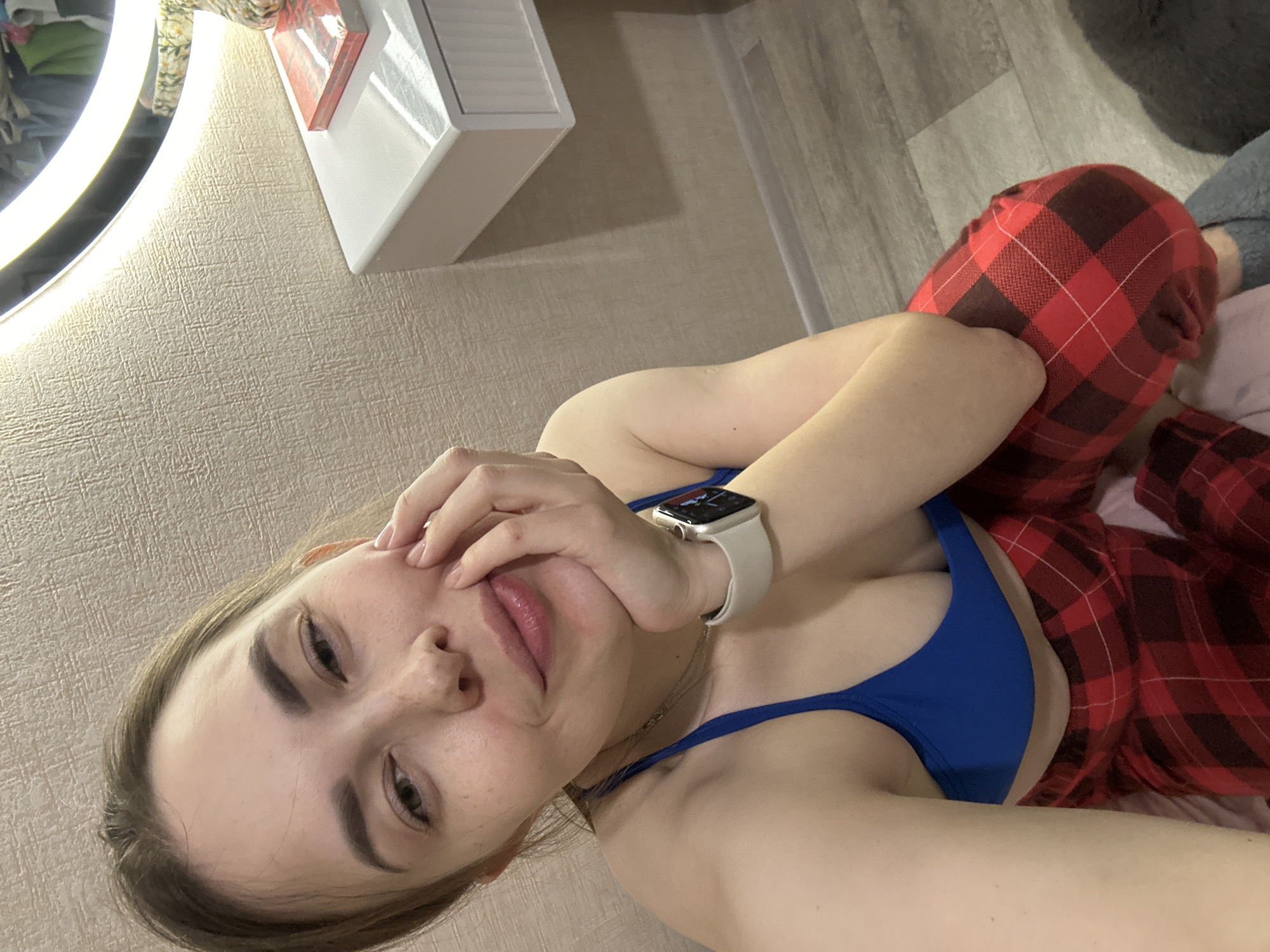 A young woman is captured in a close-up selfie, lying down in what appears to be an indoor setting, likely a bedroom or dressing area in Perm, Russia. She is looking directly at the camera with a neutral to slightly pensive expression, her lips slightly parted. Her left hand is raised to her face, subtly covering her mouth or cheek, and on her left wrist, she wears a light-colored smartwatch with a digital display.
She is dressed casually in a royal blue bra or swimsuit top and red and black plaid pajama pants. The background reveals a segment of a round mirror illuminated by a bright LED light strip on the left, casting a soft glow. To the upper right, a white dresser or cabinet unit with visible drawers is positioned against a textured, light beige or off-white wallpapered wall. Further right, a section of wooden or laminate flooring is visible. The overall scene suggests a relaxed, personal moment. No discernible text is visible in the image.