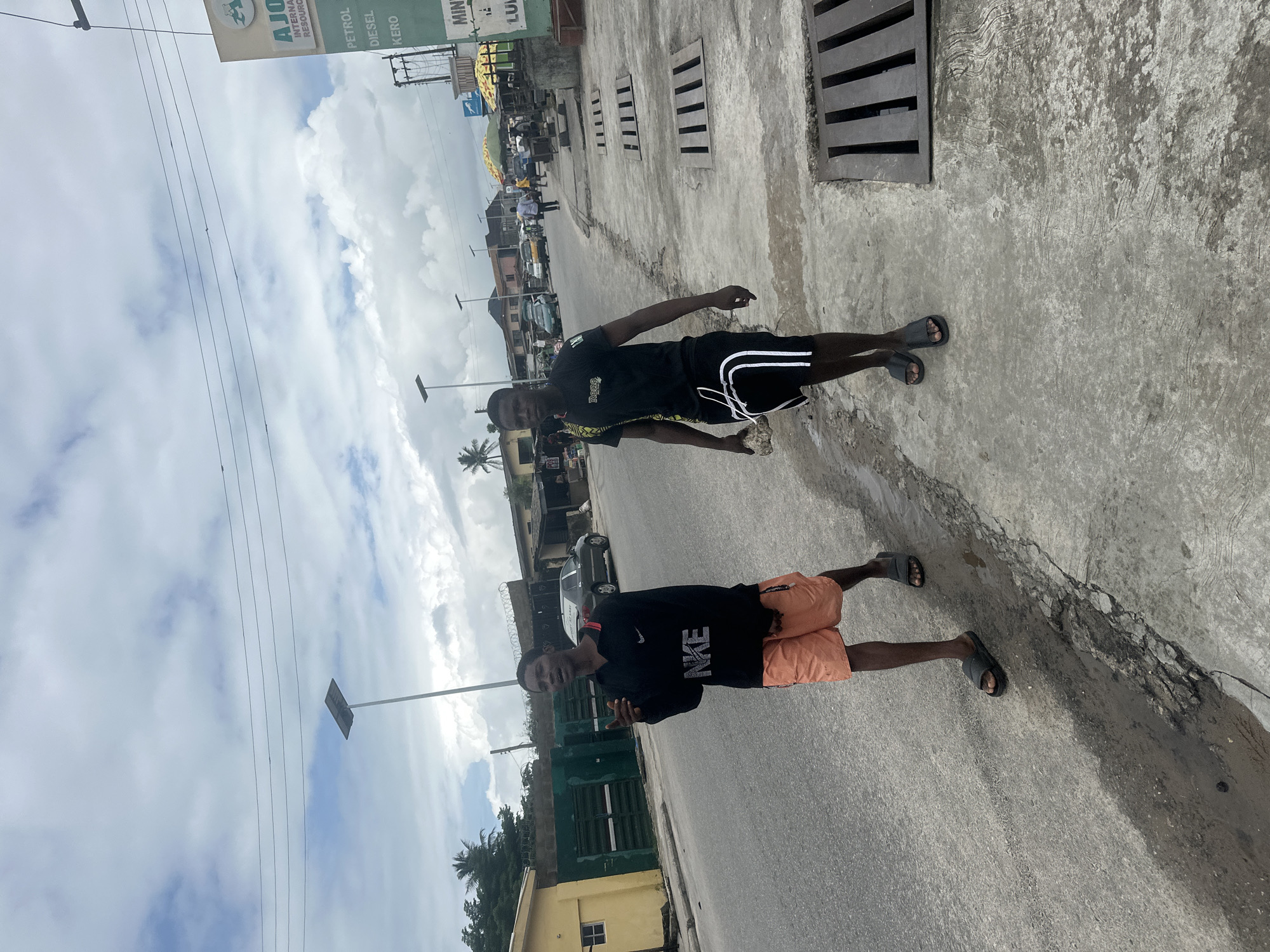 Two young men are walking on a sidewalk in Badagry, Nigeria. The man on the left is wearing a black long-sleeved shirt with "NIKE" printed on it in white, and orange shorts. He is looking at the person taking the photo and pointing. The man on the right is wearing a black t-shirt and black shorts with white stripes. He is looking forward and carrying a rock in his left hand. They are walking on a concrete sidewalk next to a street. In the background, there are buildings, a sign that says "AJOG INTERNATIONAL RESOURCES" and lists prices for petrol, diesel, and kerosene, as well as parked cars and people. The sky is cloudy.