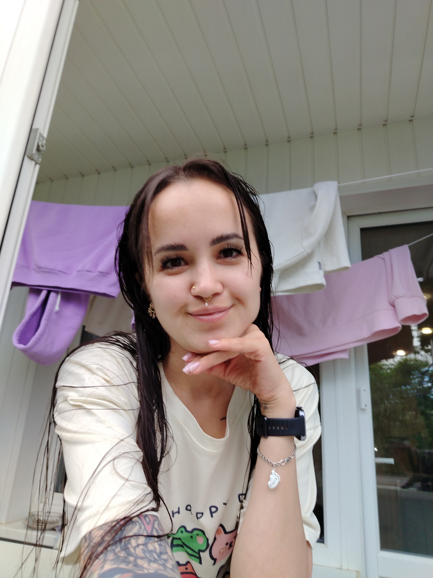 In Chaikovsky, Russia, a young woman with dark, wet hair and a nose ring is captured in a selfie. She is wearing a light-colored t-shirt with a cartoon graphic of frogs. Her right hand rests on her chin, and she sports a black smartwatch on her left wrist, adorned with a heart-shaped charm. Behind her, on a clothesline, are several pieces of clothing: a lavender hoodie, a white top, and a pink sweatshirt, suggesting a domestic setting, possibly a balcony or an outdoor laundry area. The background shows a white, paneled ceiling and a window with a reflection of trees.