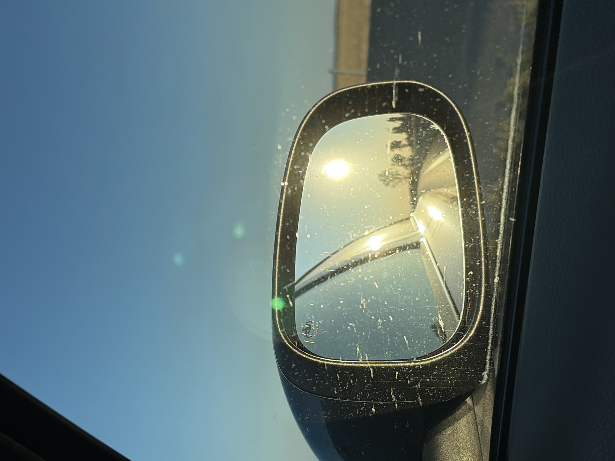 This is a close-up, medium shot of a car's side mirror reflecting a sunlit road with a line of trees and a clear blue sky. The mirror itself is speckled with dirt or water spots, and a small green light is visible on the edge of the mirror housing. The image appears to have been taken from inside the car, looking out the window. The sun's glare creates bright highlights on the mirror's surface. There is no specific indication of the location within Dolná Streda, Slovakia.