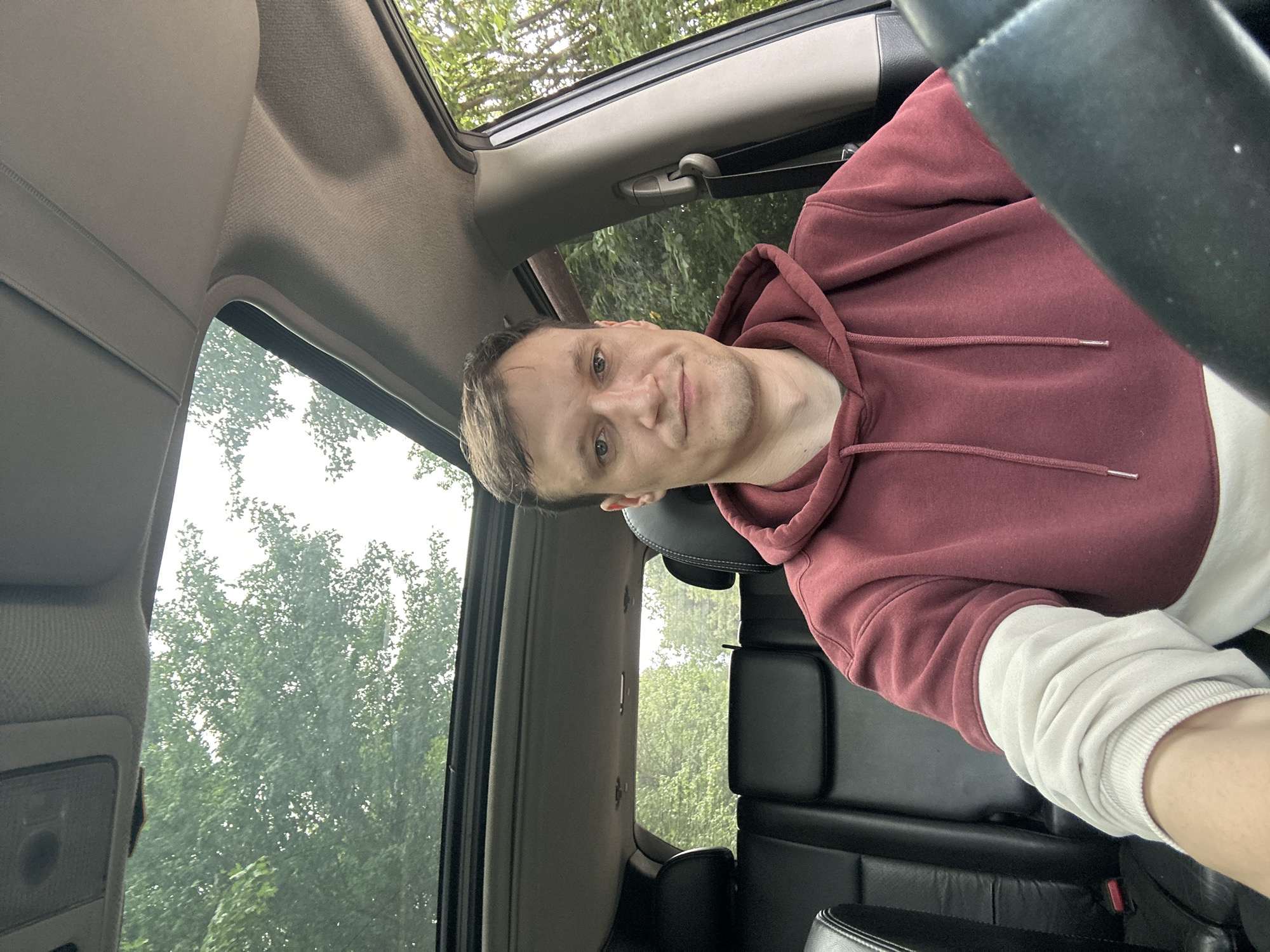 A man is sitting in the driver's seat of a car, looking at the camera. He is wearing a maroon hoodie with a white layer underneath. The car has a sunroof open, and trees can be seen through it and through the side windows. The interior of the car includes black leather seats and part of the steering wheel is visible. The scene appears to be located in a wooded area, possibly on the outskirts of the city of Korchivka, Ukraine.